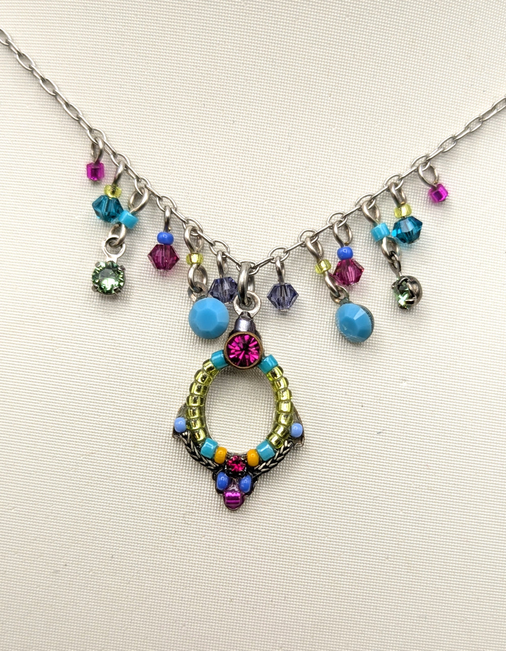 A silver-toned charm necklace displayed on a textured off-white fabric background. The necklace features a central oval pendant decorated with pink, blue, yellow, and purple beads, along with dangling colorful faceted beads in pink, blue, green, and purple.