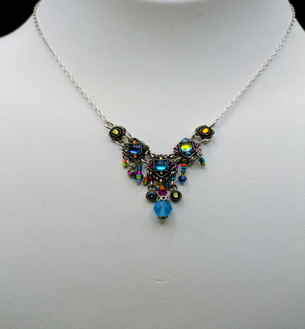 A silver-toned statement necklace displayed on a white mannequin bust. The necklace features a central cluster of colorful iridescent and blue beads, small dangling charms, and a faceted light blue bead at the bottom, suspended from a delicate chain.