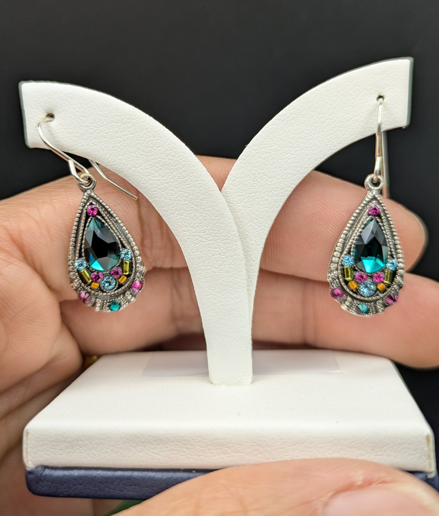 Teardrop-shaped earrings with colorful gemstones on a white stand held by a hand against a black background.