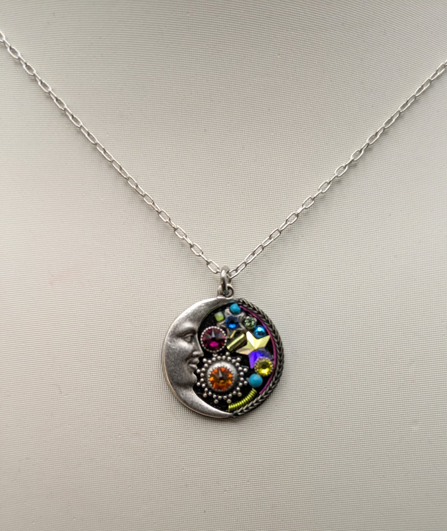 A silver pendant necklace featuring a crescent moon design on one side and a colorful, gemstone-encrusted face on the other, displayed against a plain light gray background.