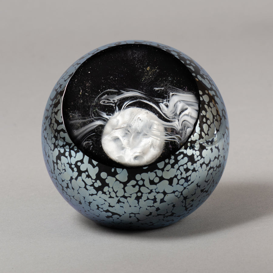 A glass paperweight featuring a stylized full moon and wind swept clouds design, with a black and blue color pattern.