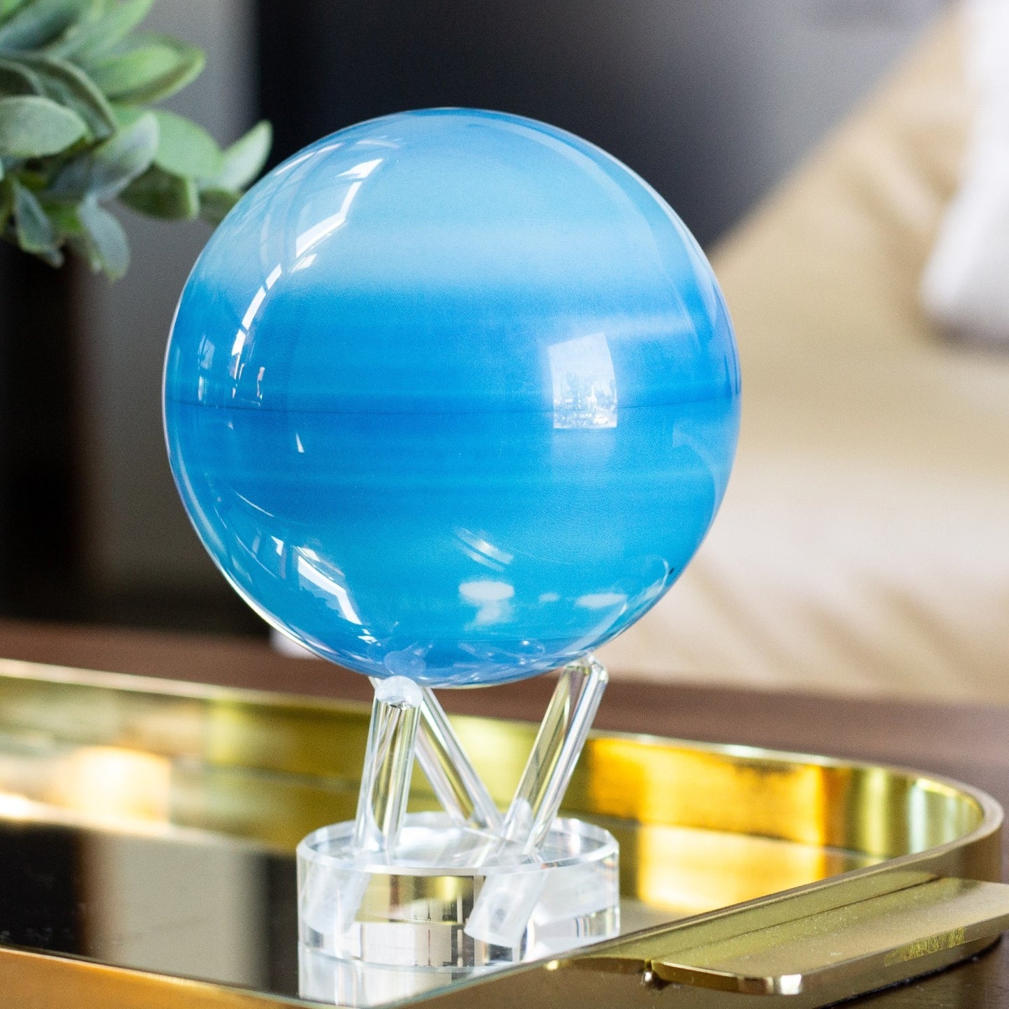 A blue spherical MOVA globe on a clear stand, displaying the icy tones of Uranus, placed on a gold surface.