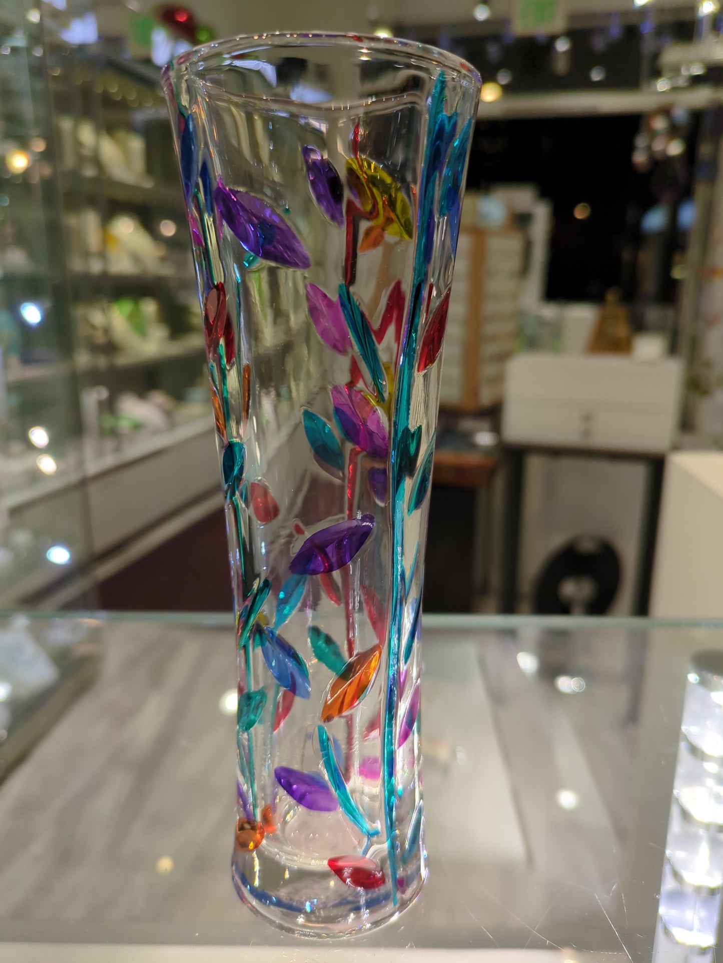 A multicolored glass vase with a floral Tree of Life design, displayed in a store with various decorative items around it.