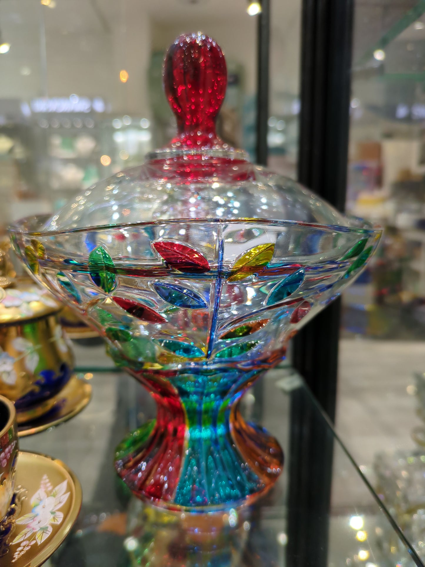 A multicolored glass dish with lid resembling a tree of life, displayed on a shelf with other glass items.