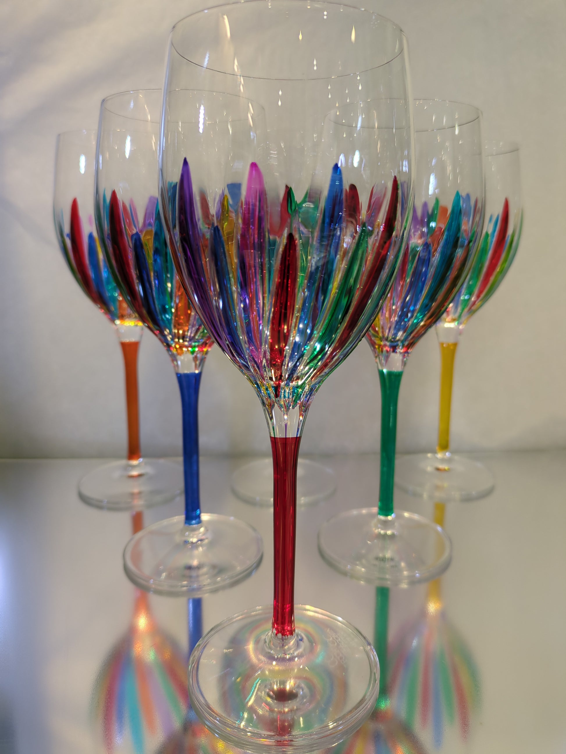 A set of multicolored wine glasses with a clear upper part and colorful lower part, with a reflective surface below.