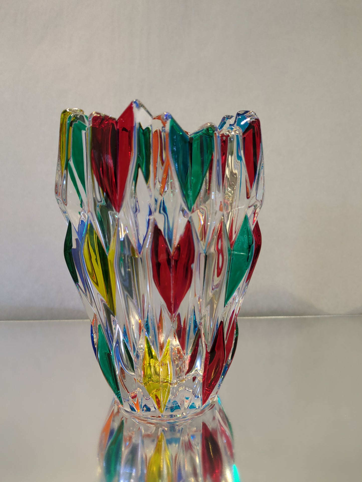 A multicolored glass vase with a geometric pattern, featuring colors such as red, green, and yellow, standing on a reflective surface.