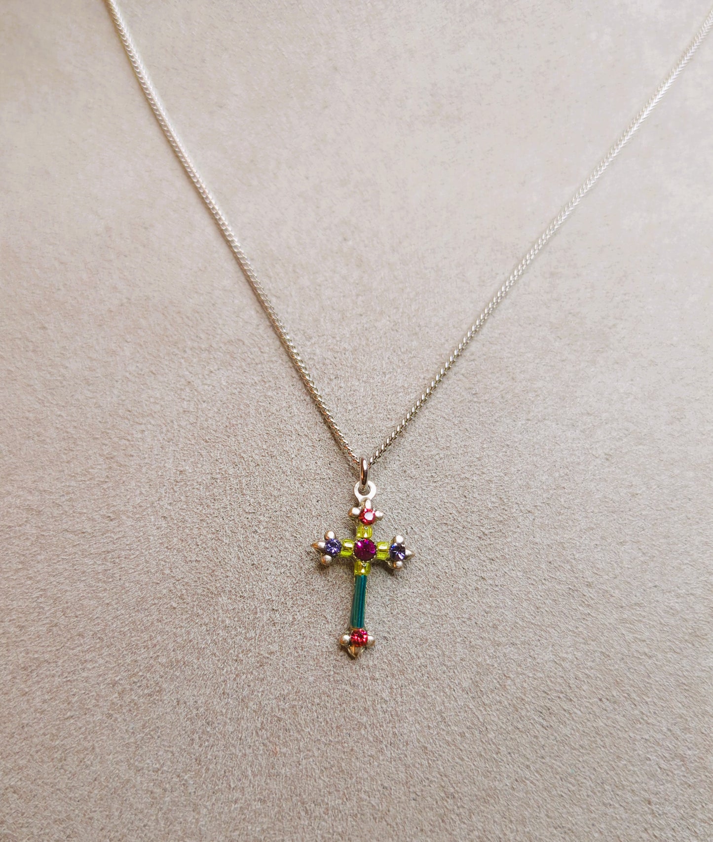 A multicolored cross necklace with a chain link and a lobster claw clasp on gray pad.