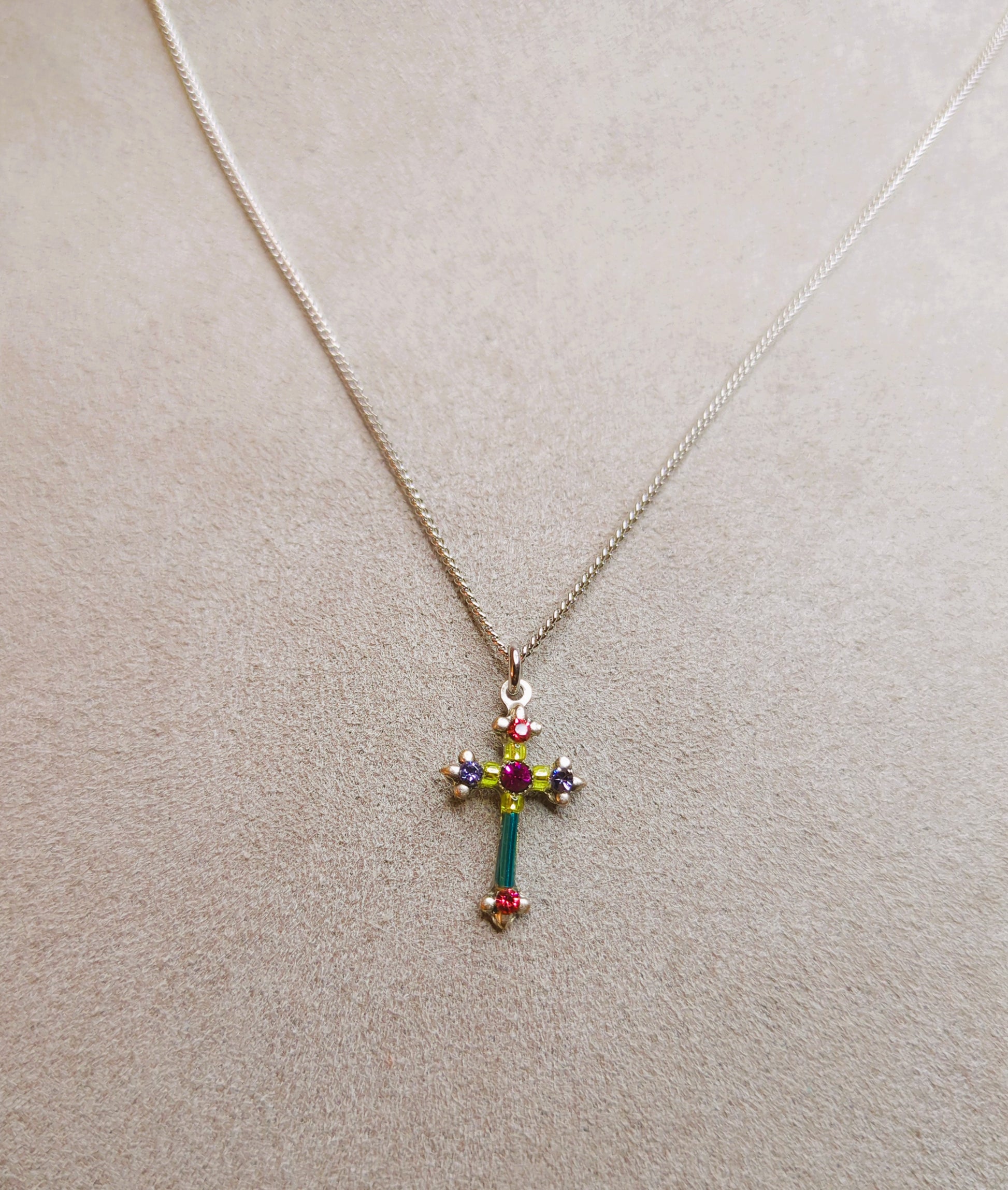 A multicolored cross necklace with a chain link and a lobster claw clasp on gray pad.