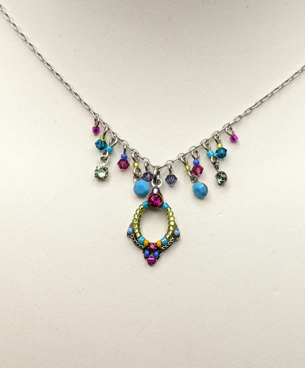 A silver-toned necklace displayed on a white mannequin bust, featuring a central gold-toned circular pendant embellished with multicolored rhinestones and several dangling colorful beads.