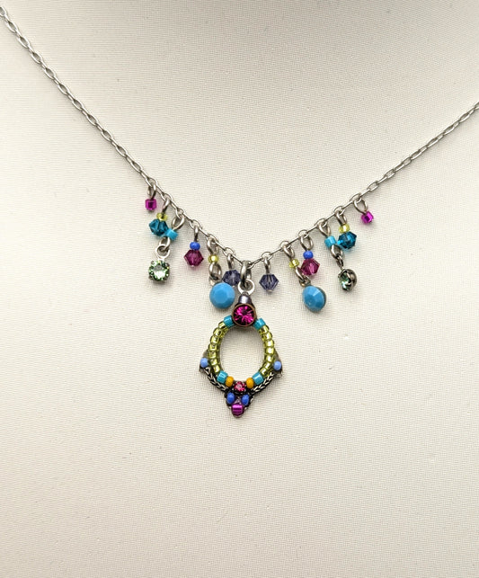 A silver-toned necklace displayed on a white mannequin bust, featuring a central gold-toned circular pendant embellished with multicolored rhinestones and several dangling colorful beads.