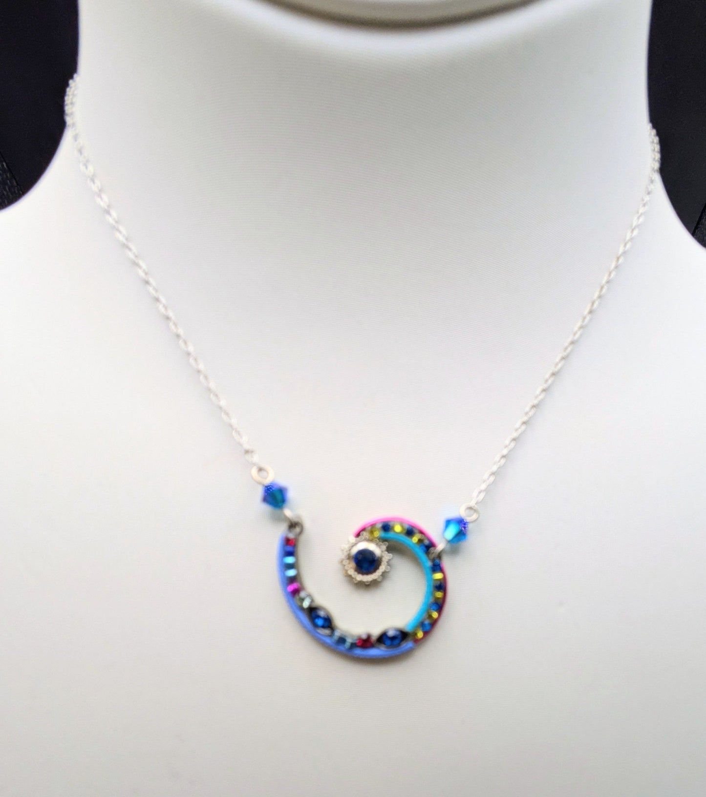 A silver chain necklace with a spiral pendant featuring blue, red, and yellow beads, displayed on a white mannequin bust.