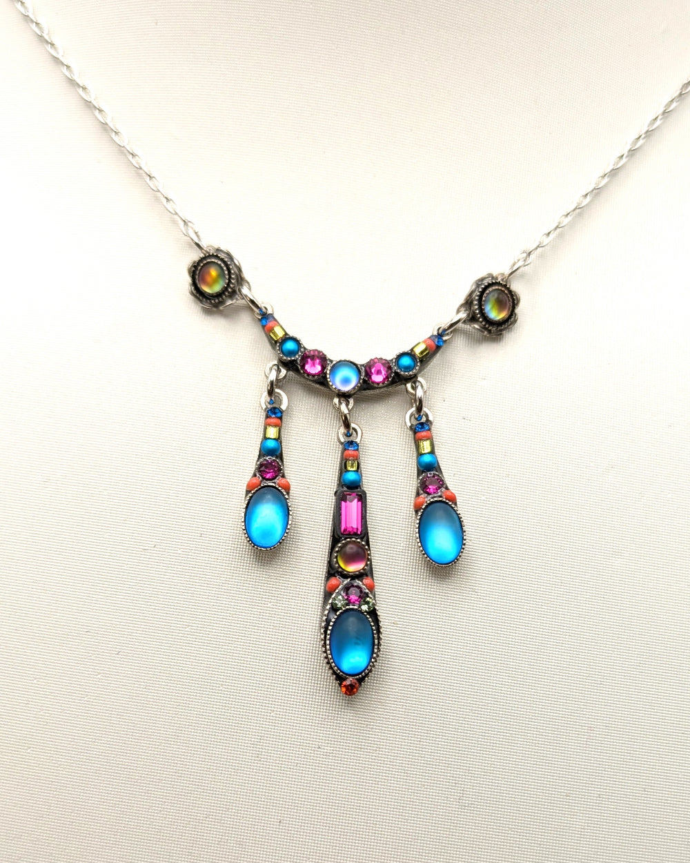 A silver-toned necklace with a decorative pendant featuring bright blue, pink, and iridescent gemstones, displayed on a light beige background.