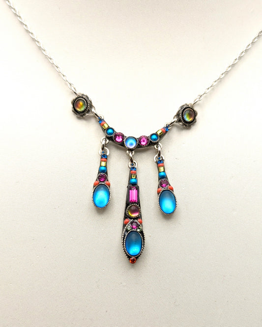 A silver-toned necklace with a decorative pendant featuring bright blue, pink, and iridescent gemstones, displayed on a light beige background.