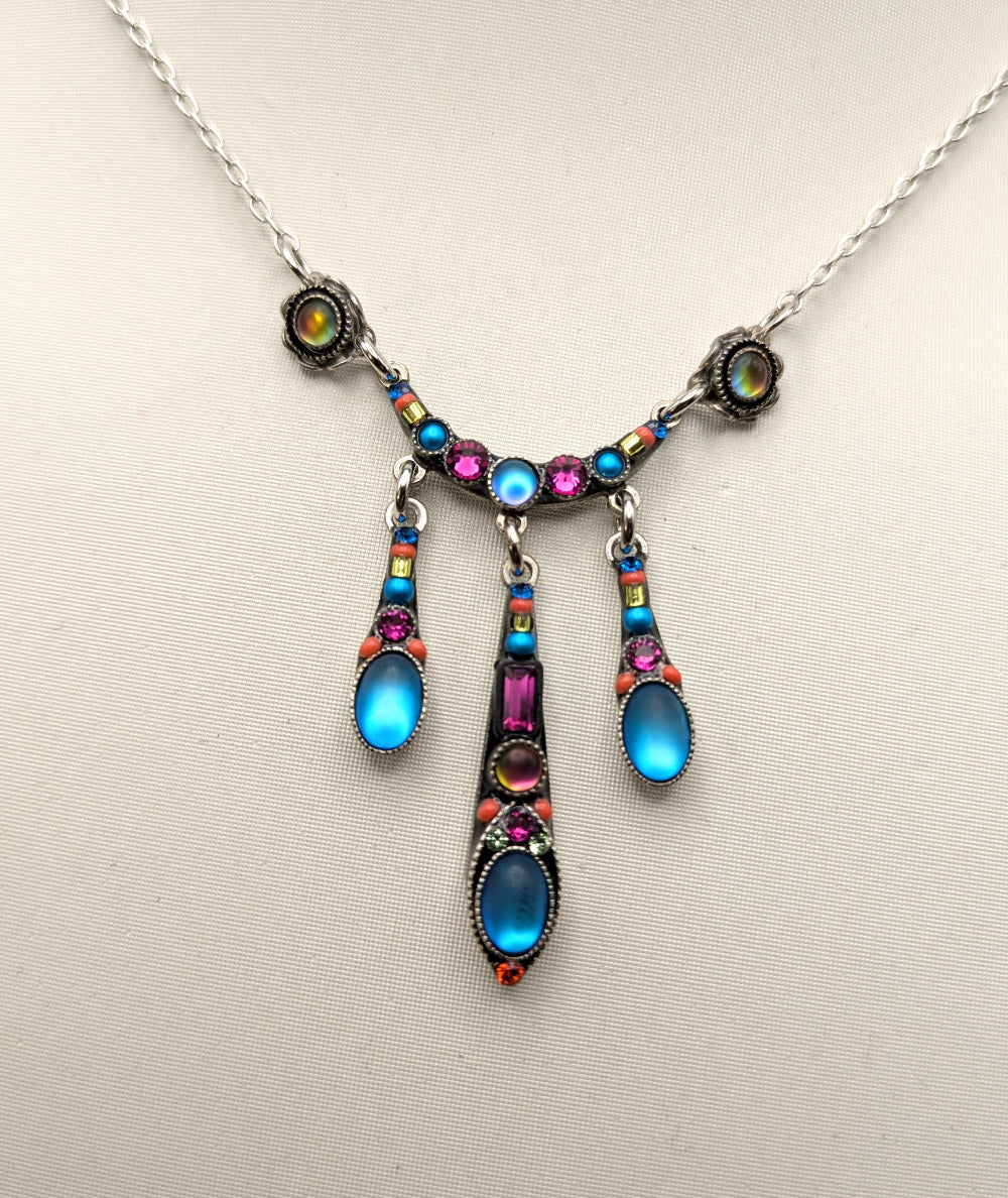 A silver-toned pendant necklace with vibrant, multicolored rhinestone accents and three teardrop-shaped blue cabochon drops, displayed on a neutral beige background.