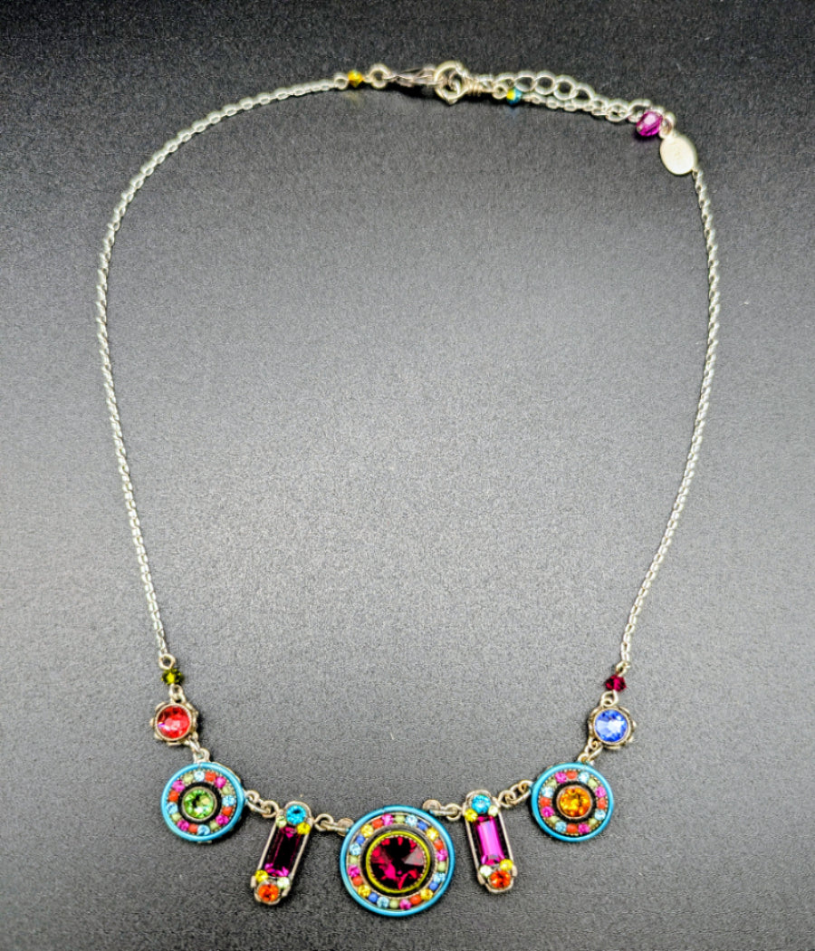 A silver-toned chain necklace with a colorful statement bib-style pendant. The necklace features five circular and rectangular charms in vibrant turquoise, pink, orange, yellow, green, and purple, arranged on a dark gray textured background.