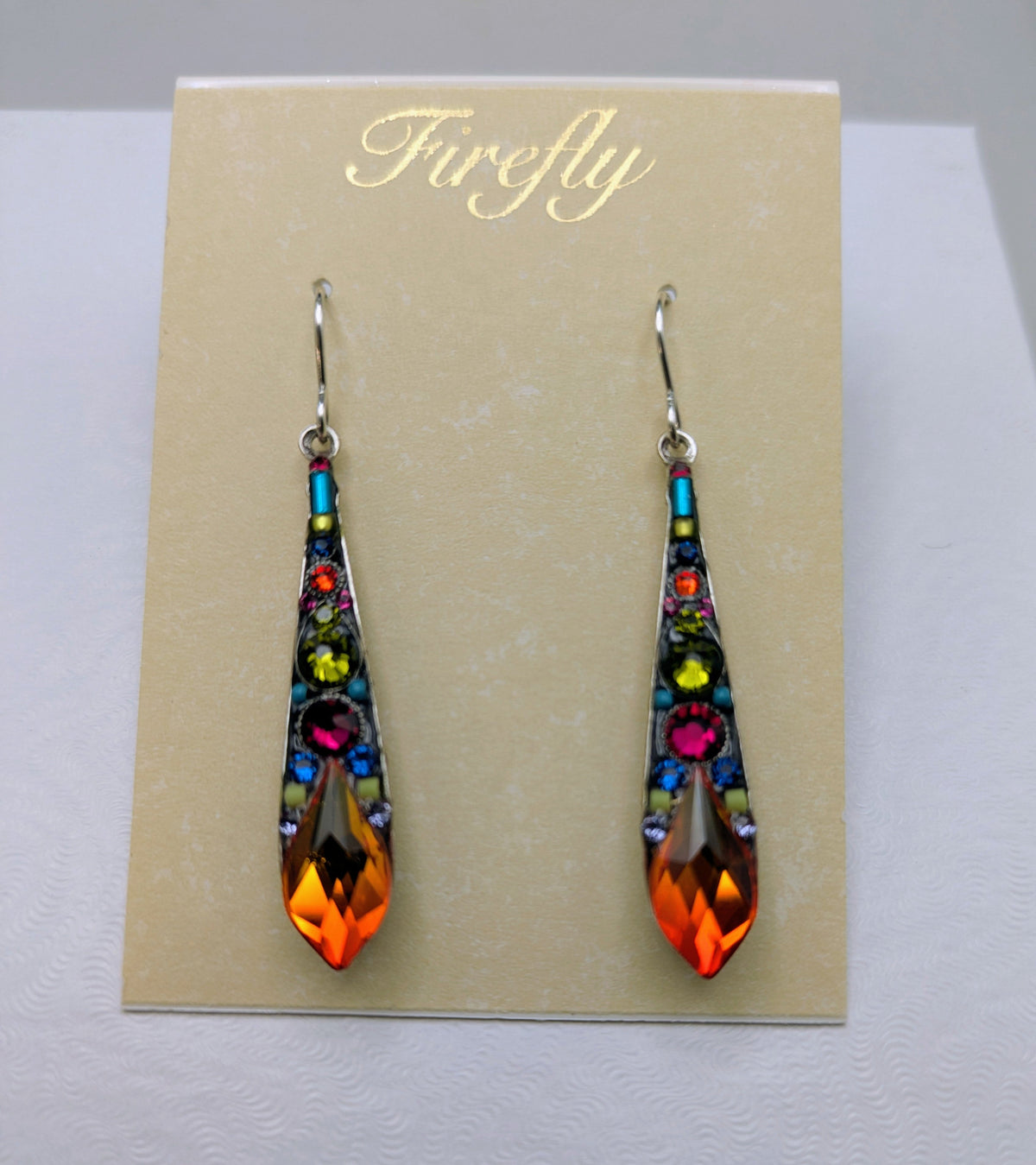 A pair of long teardrop-shaped dangling earrings displayed on a beige card labeled 'Firefly.' The earrings have silver hooks and silver-toned settings with multiple colorful rhinestones in blue, green, yellow, pink, and orange, plus a large faceted orange crystal at the bottom.