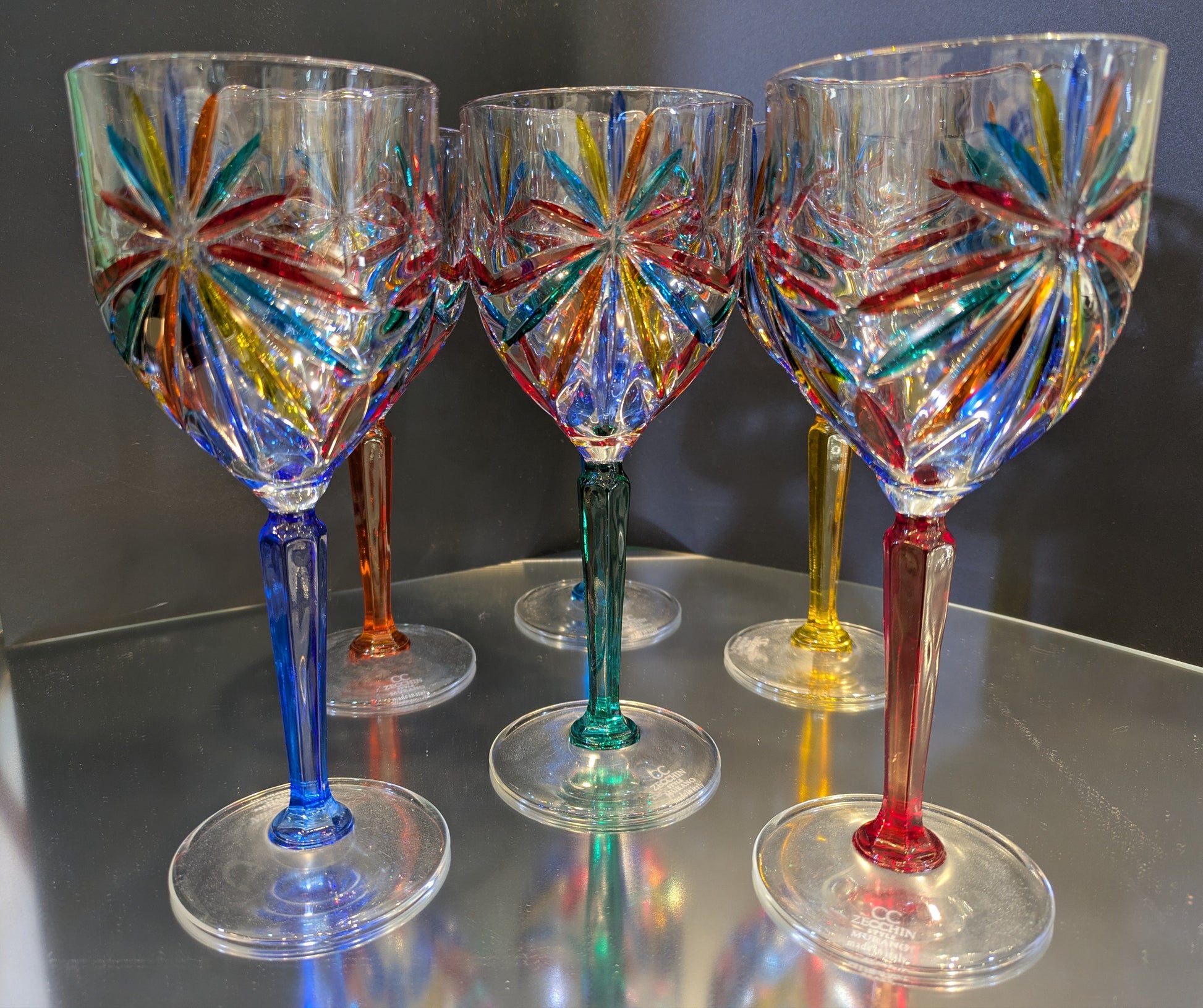 A set of six multicolored wine glasses with a geometric pattern on a reflective surface.