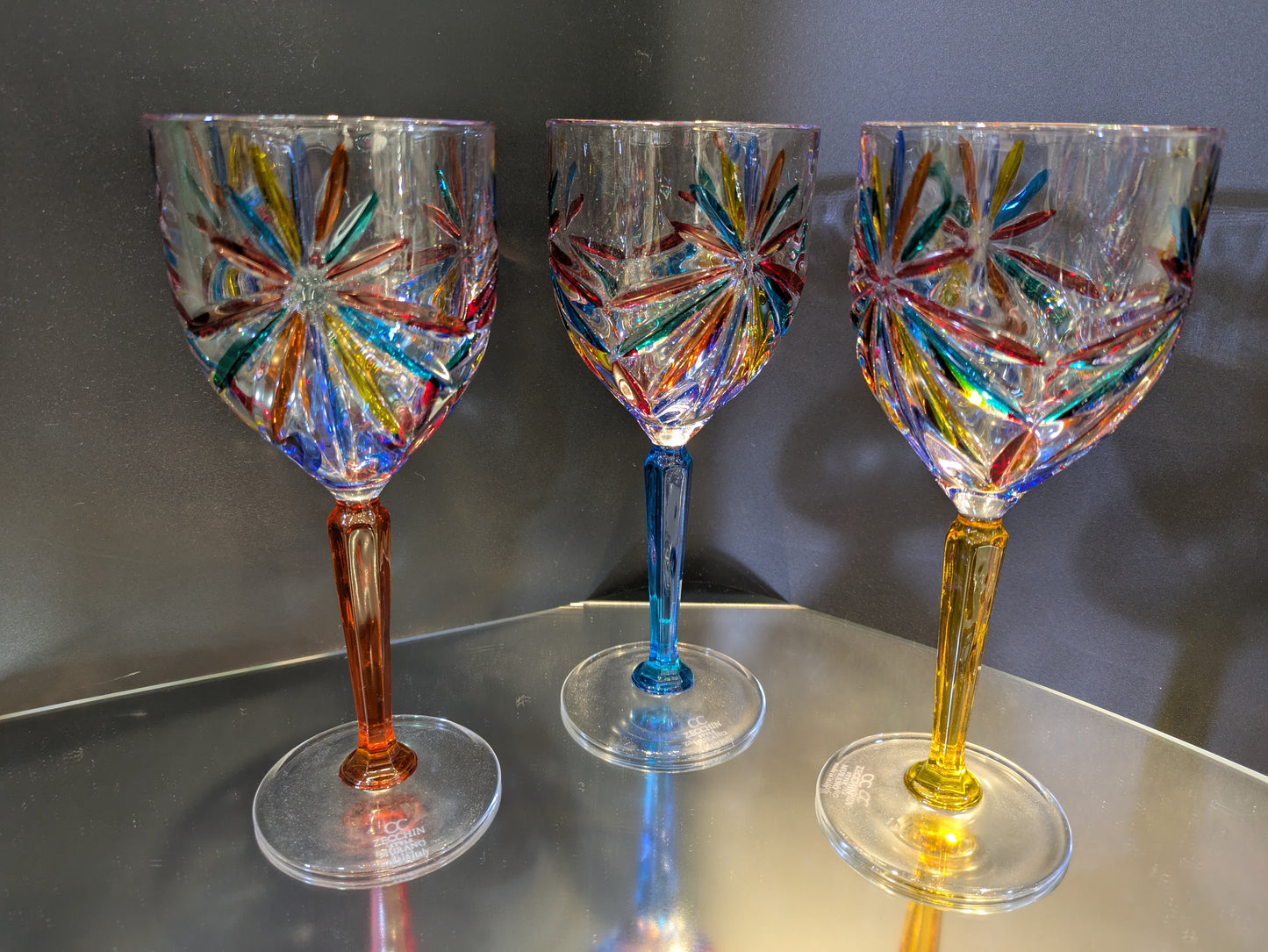 A set of three multicolored wine glasses with a geometric pattern on a reflective surface.