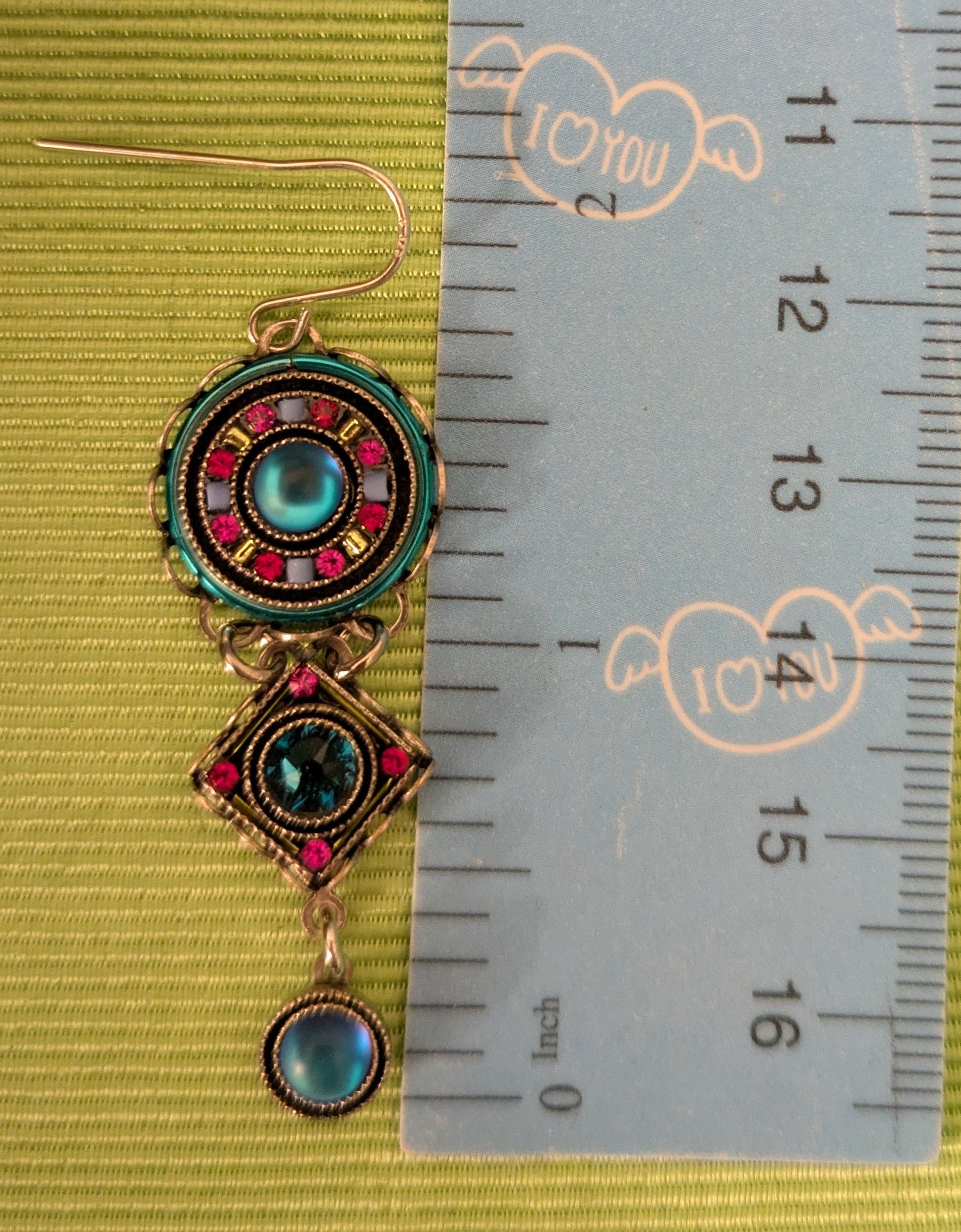 Pair of decorative earrings on a ruler for scale against a green background