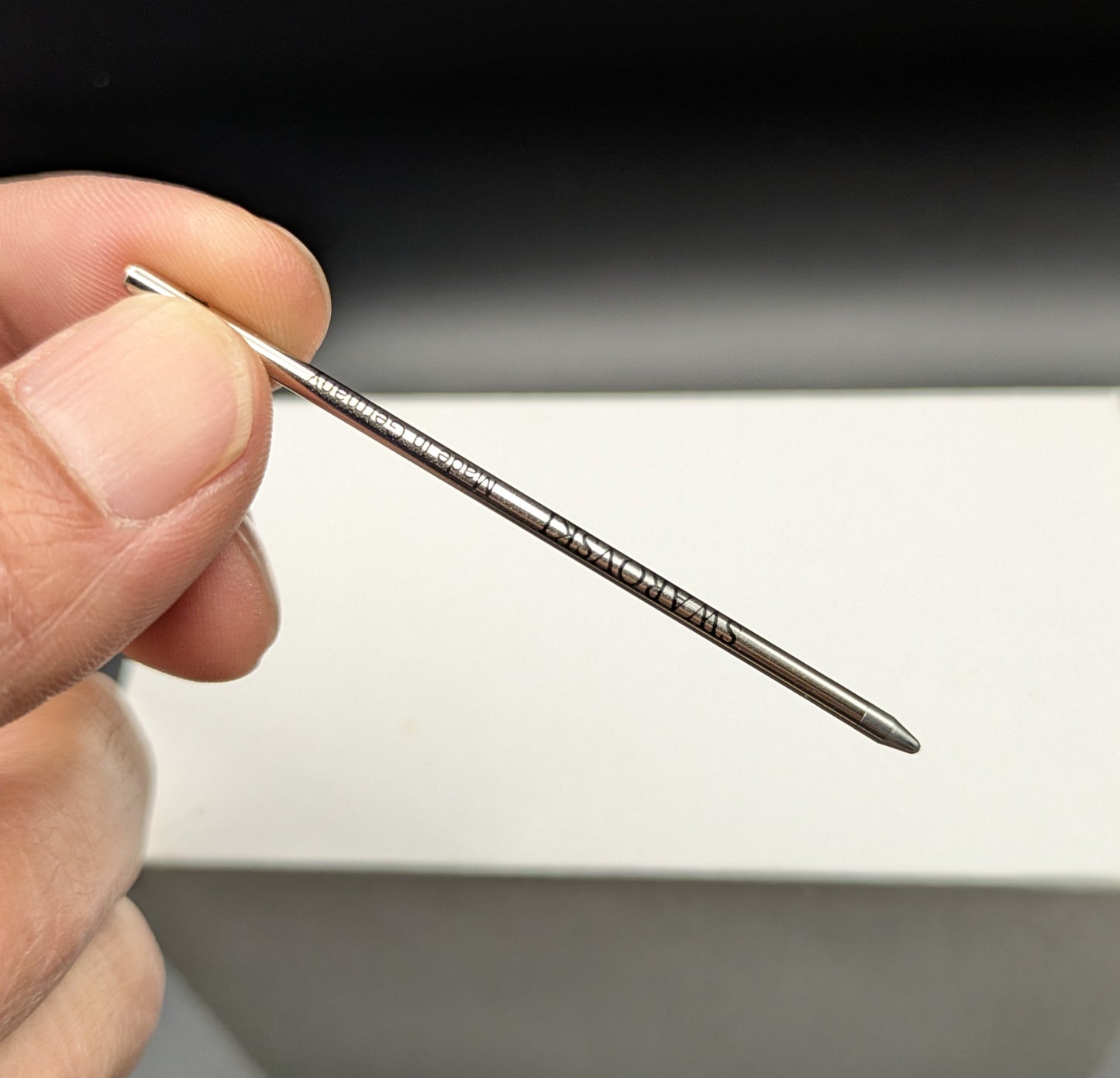 Hand holding a pen refill in front of a white box on a dark background
