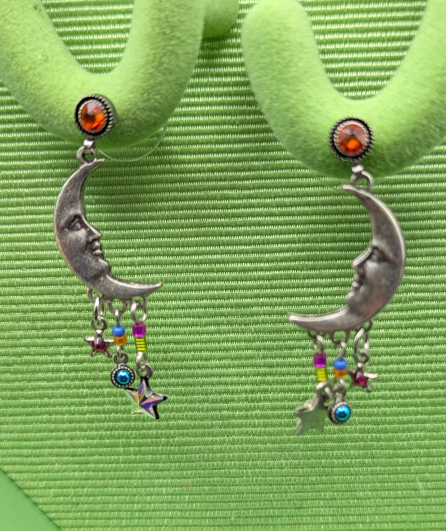 Silver moon-shaped earrings with colorful charms on a green textured background
