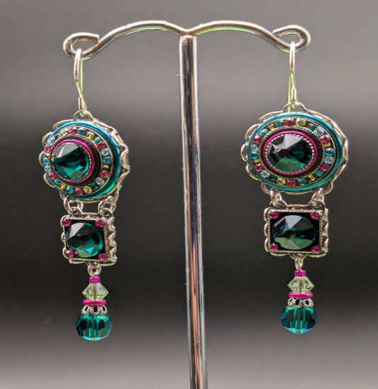 Pair of ornate earrings with green and pink gemstones on a stand against a gray background.