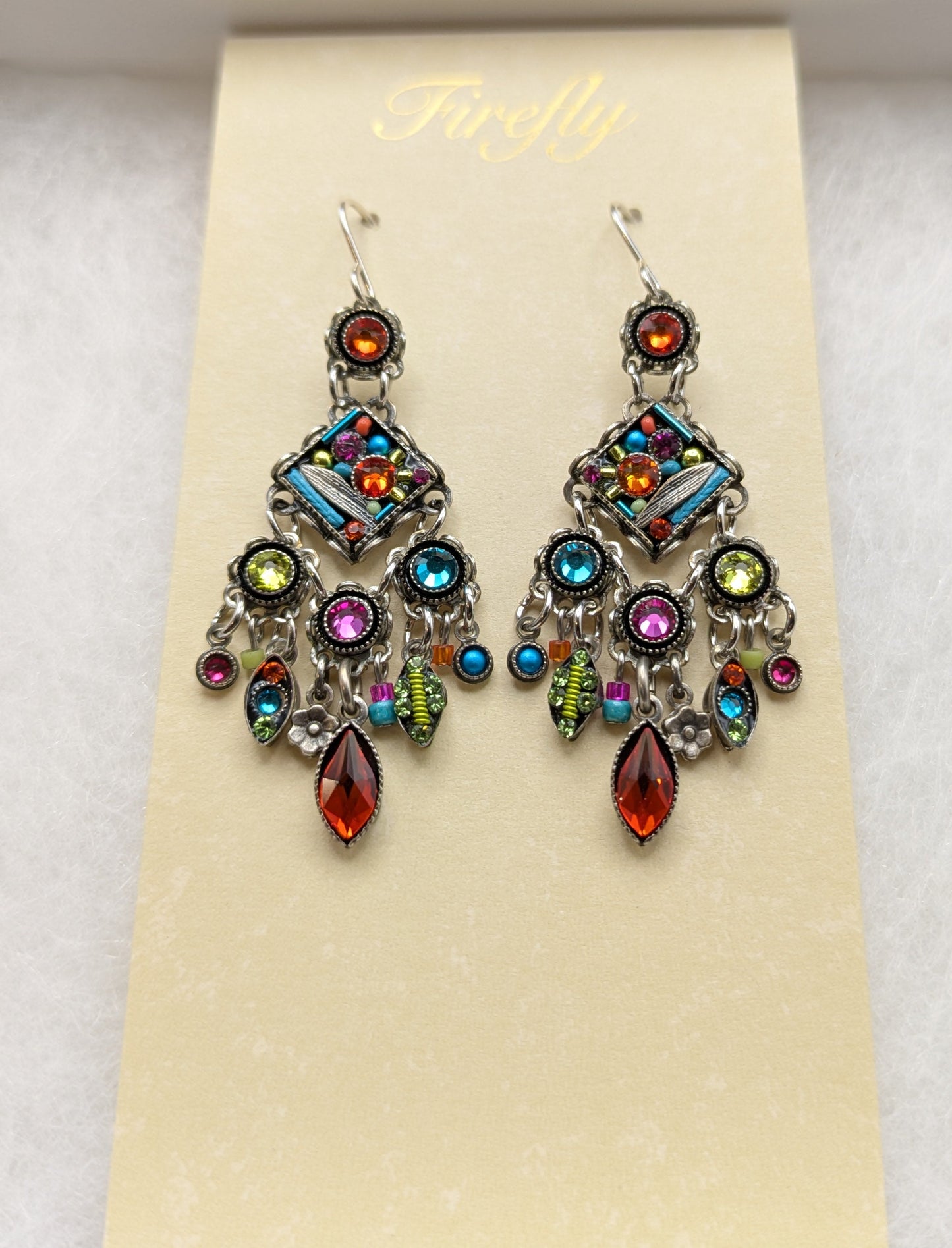 A pair of colorful dangling earrings displayed on a cream card inside a white jewelry box. The earrings feature silver-toned metal with multiple vibrant gemstones in blue, green, pink, orange, and yellow, along with teardrop-shaped red-orange crystals at the bottom.