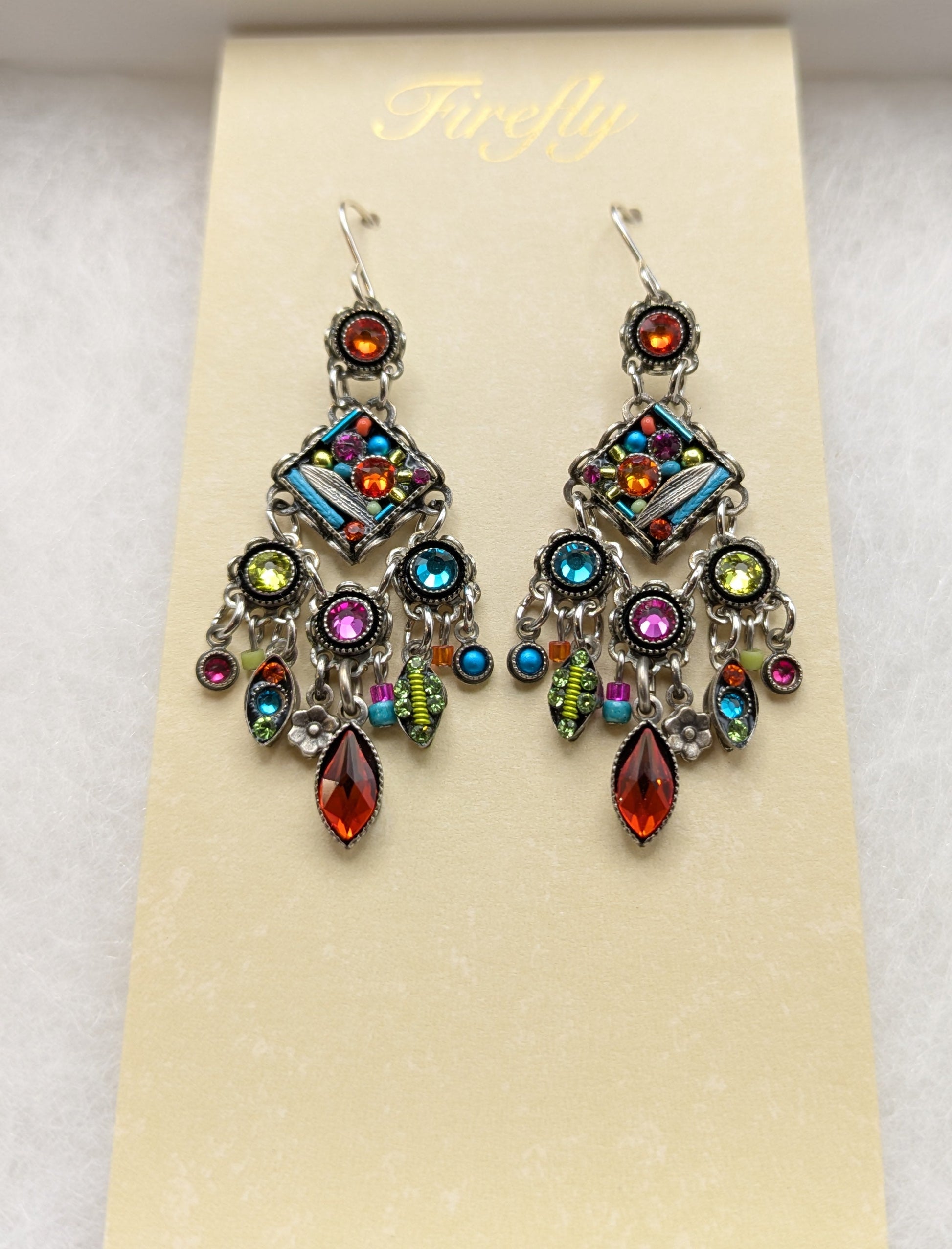 A pair of colorful dangling earrings displayed on a cream card inside a white jewelry box. The earrings feature silver-toned metal with multiple vibrant gemstones in blue, green, pink, orange, and yellow, along with teardrop-shaped red-orange crystals at the bottom.