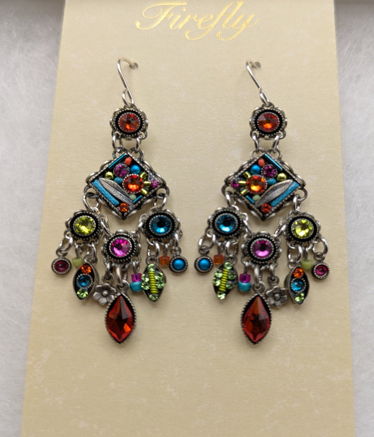 A pair of ornate dangling earrings displayed on a cream card labeled 'Firefly.' The earrings have silver-toned metal settings with multiple colorful rhinestones and teardrop charms in red, blue, green, pink, and purple tones.