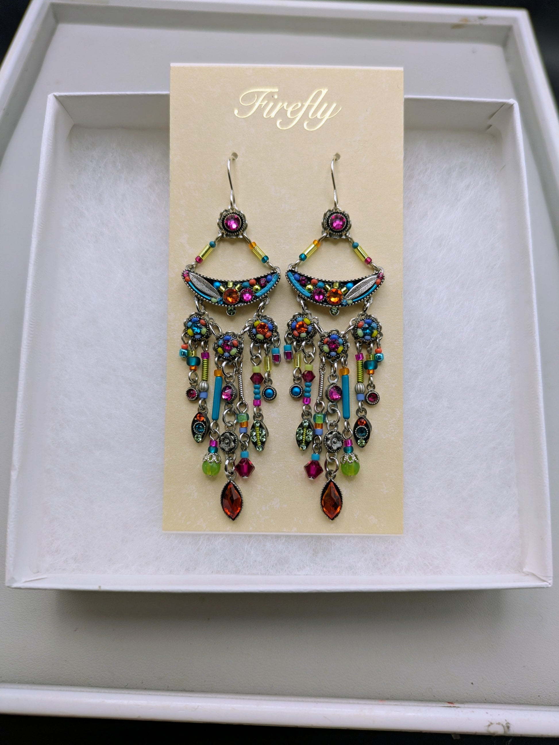 A pair of colorful dangling earrings displayed on a cream card labeled 'Firefly' inside a white gift box. The earrings feature silver-toned metal, turquoise blue enamel, and multiple dangling multicolored stones.