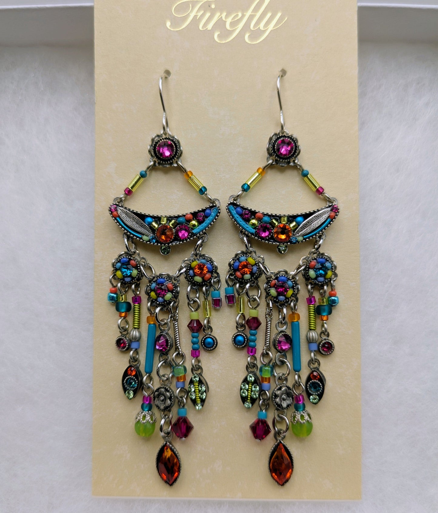 A pair of ornate dangling earrings displayed on a cream card labeled 'Firefly' inside a white box. The earrings feature silver-toned metal, multiple dangling bead and crystal chains.