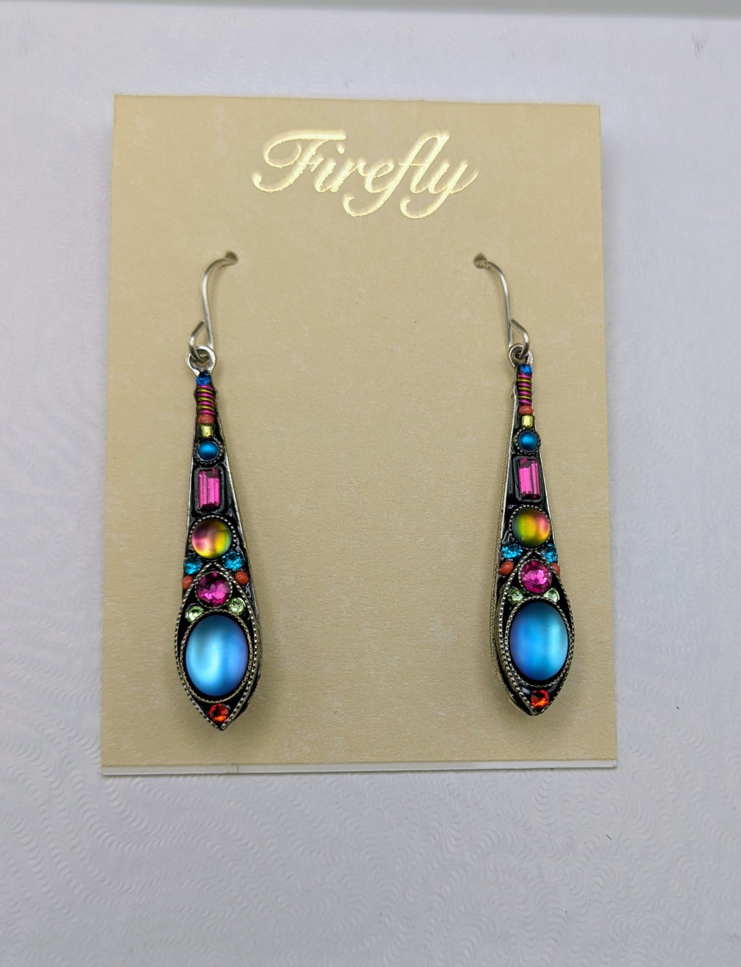 A pair of ornate drop earrings displayed on a beige card labeled 'Firefly.' The earrings have silver-toned hooks and dark metal settings with multiple colorful stones, including blue oval centers, pink and yellow accents, and small red details.