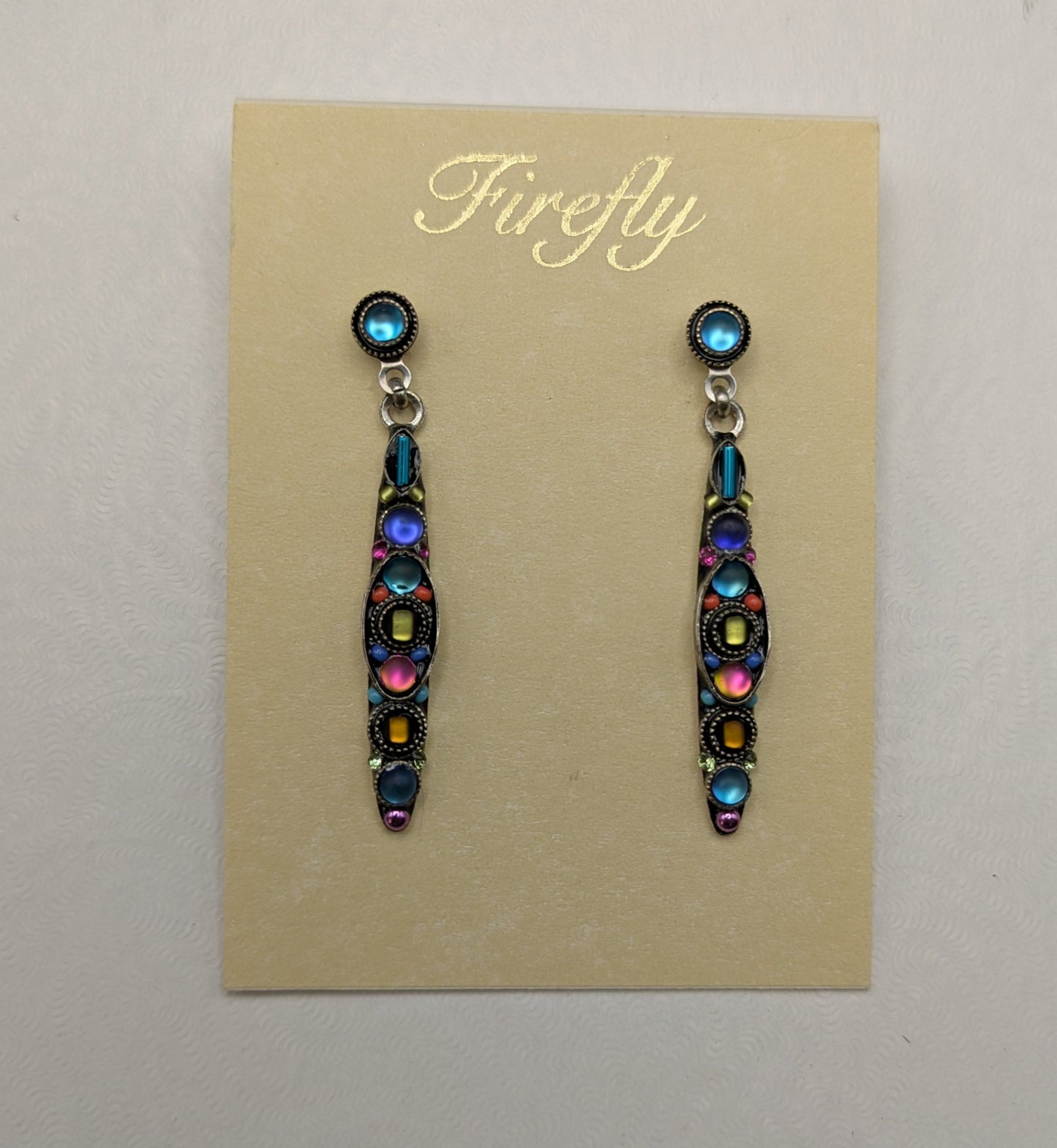 A pair of Firefly brand dangle earrings displayed on a beige card insert, set against a white gift box on a neutral gray background.