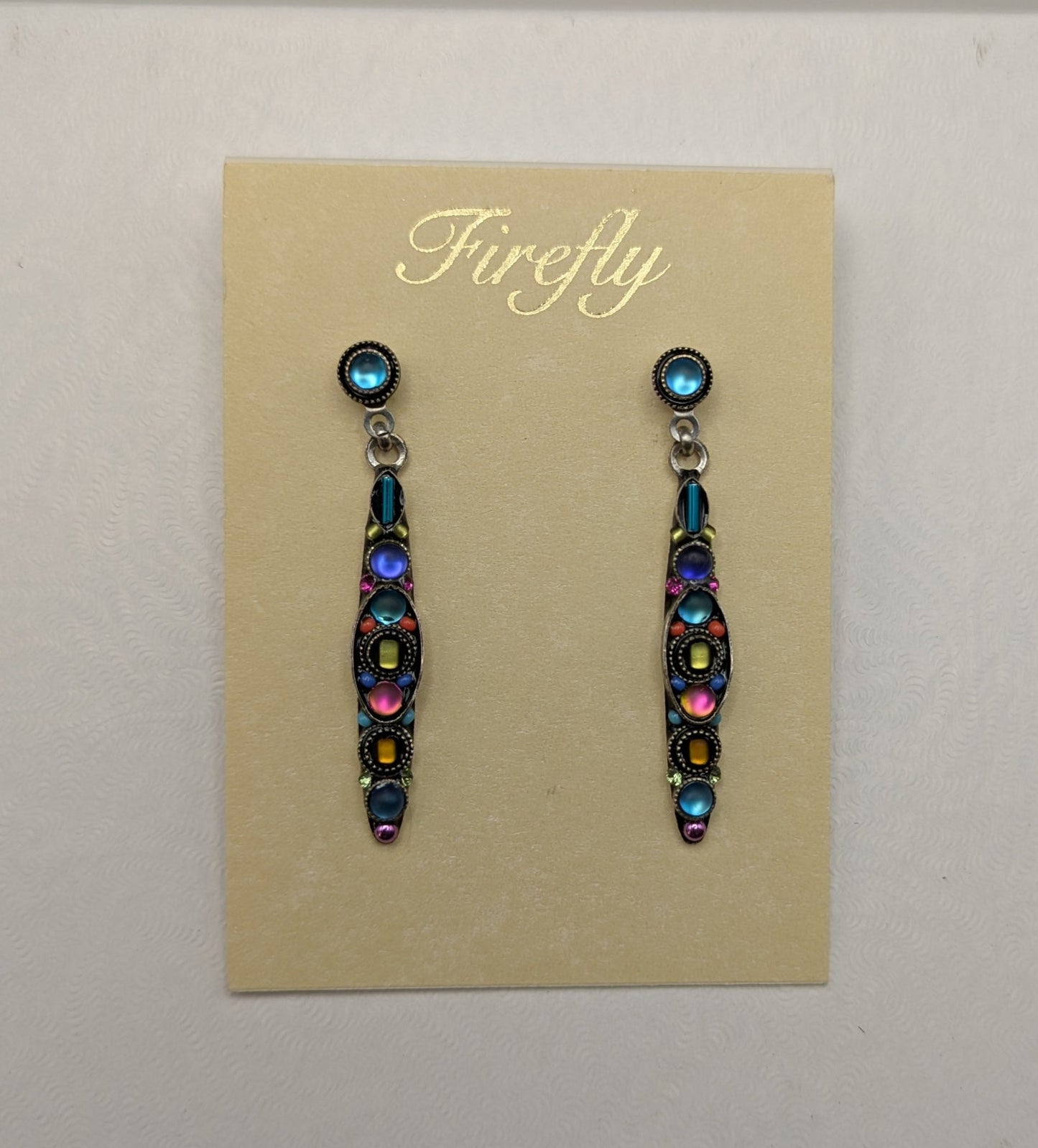 A pair of dangling earrings displayed on a beige card labeled 'Firefly,' placed on top of a white box against a light gray background.