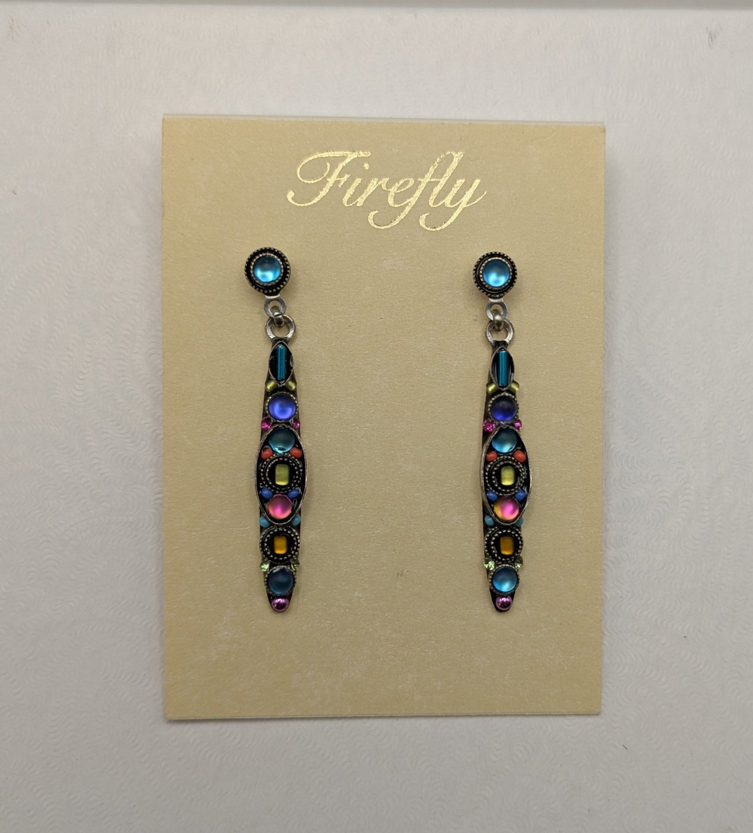 A pair of dangling earrings displayed on a beige card labeled 'Firefly,' placed on top of a white box against a light gray background.