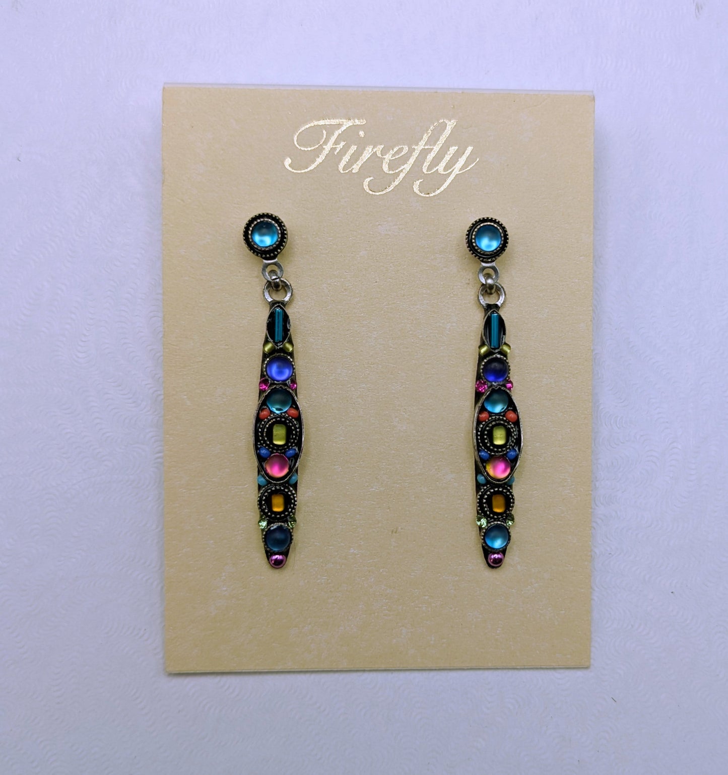 A pair of colorful drop earrings displayed on a beige card labeled 'Firefly,' placed on top of a white square box against a light gray background.