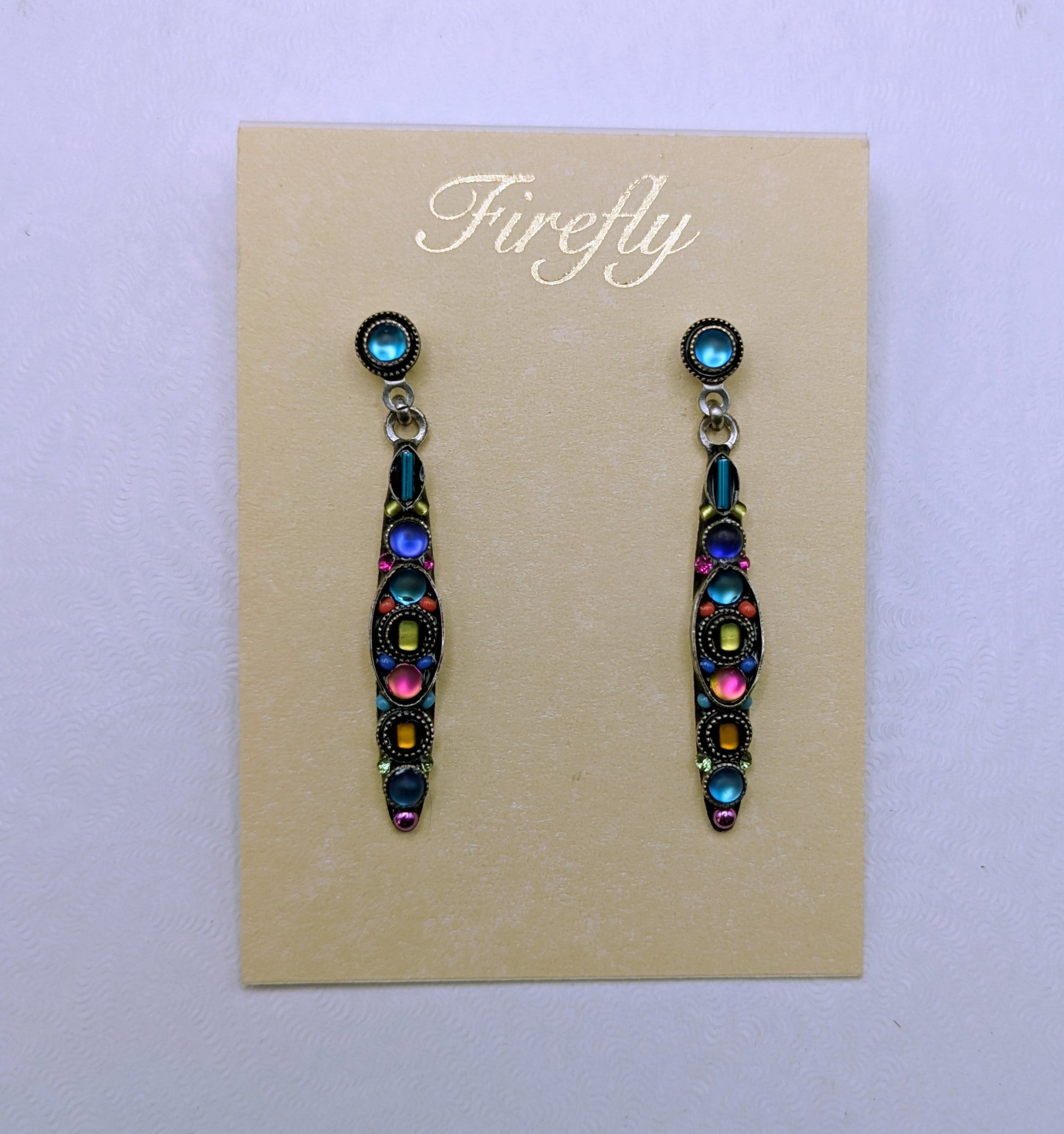 A pair of colorful drop earrings displayed on a beige card labeled 'Firefly,' placed on top of a white square box against a light gray background.