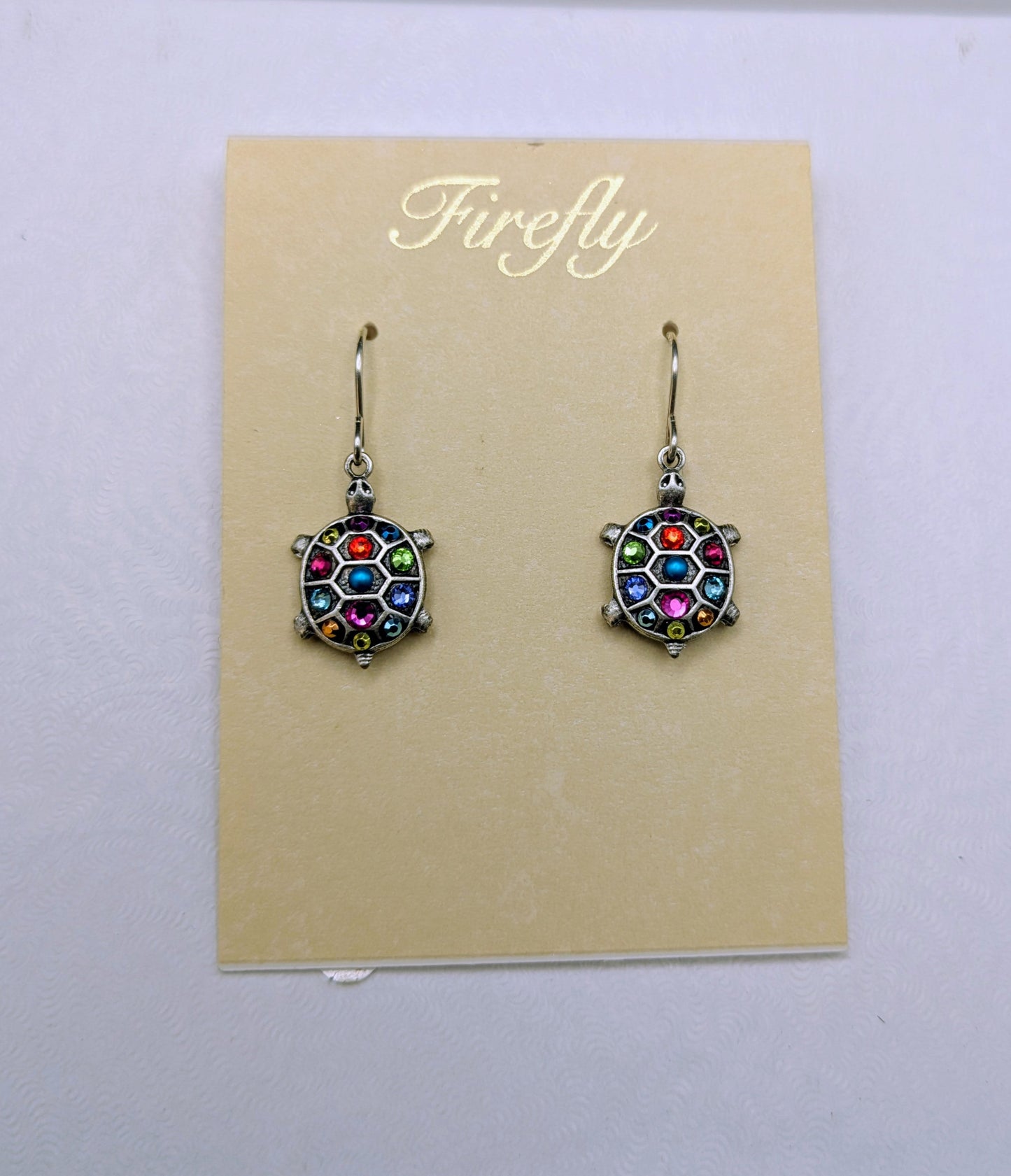 A pair of Firefly brand dangle earrings with colorful faceted crystal-like centers and silver-toned hooks, displayed on a beige card over a white box.