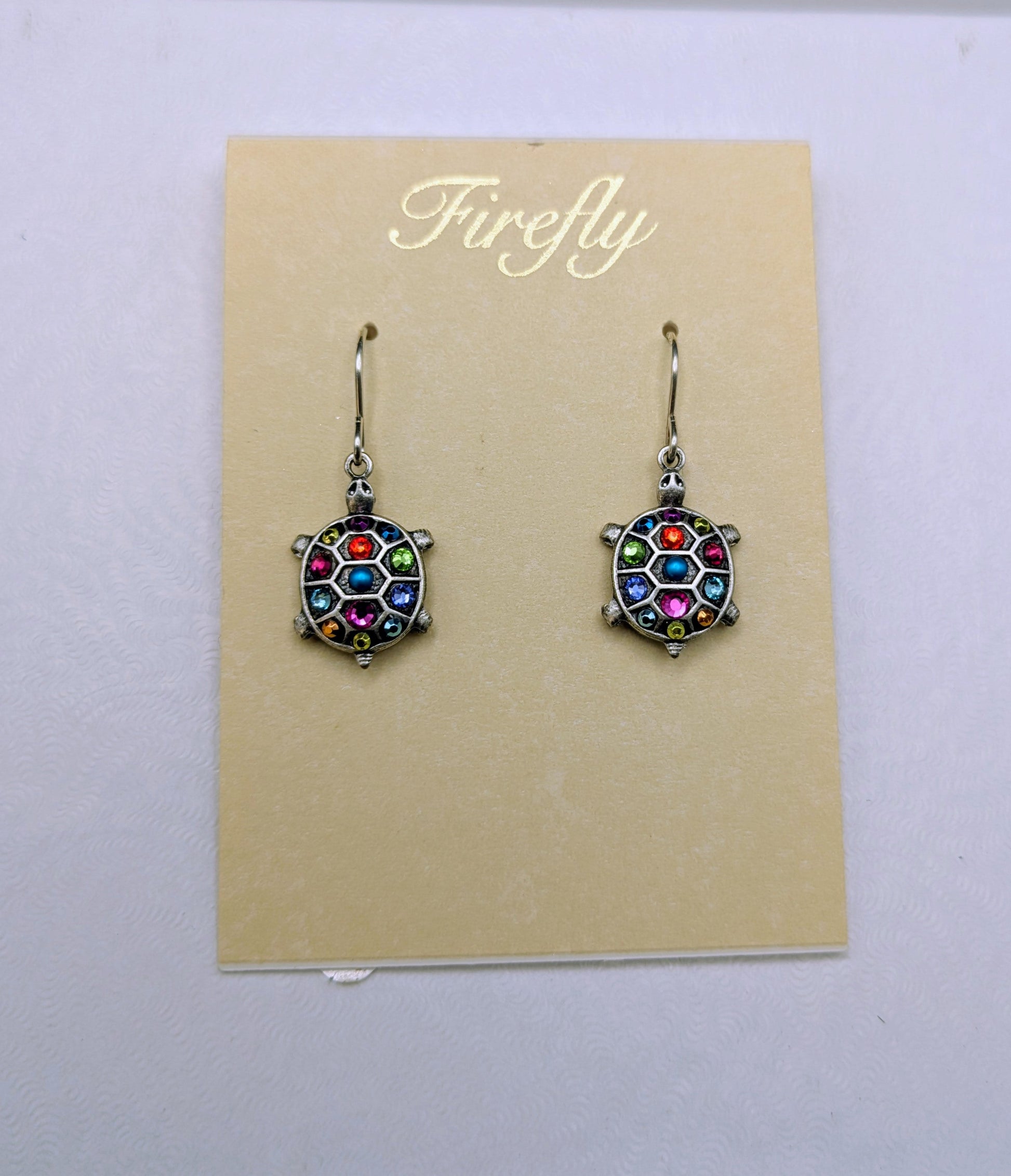 A pair of Firefly brand dangle earrings with colorful faceted crystal-like centers and silver-toned hooks, displayed on a beige card over a white box.