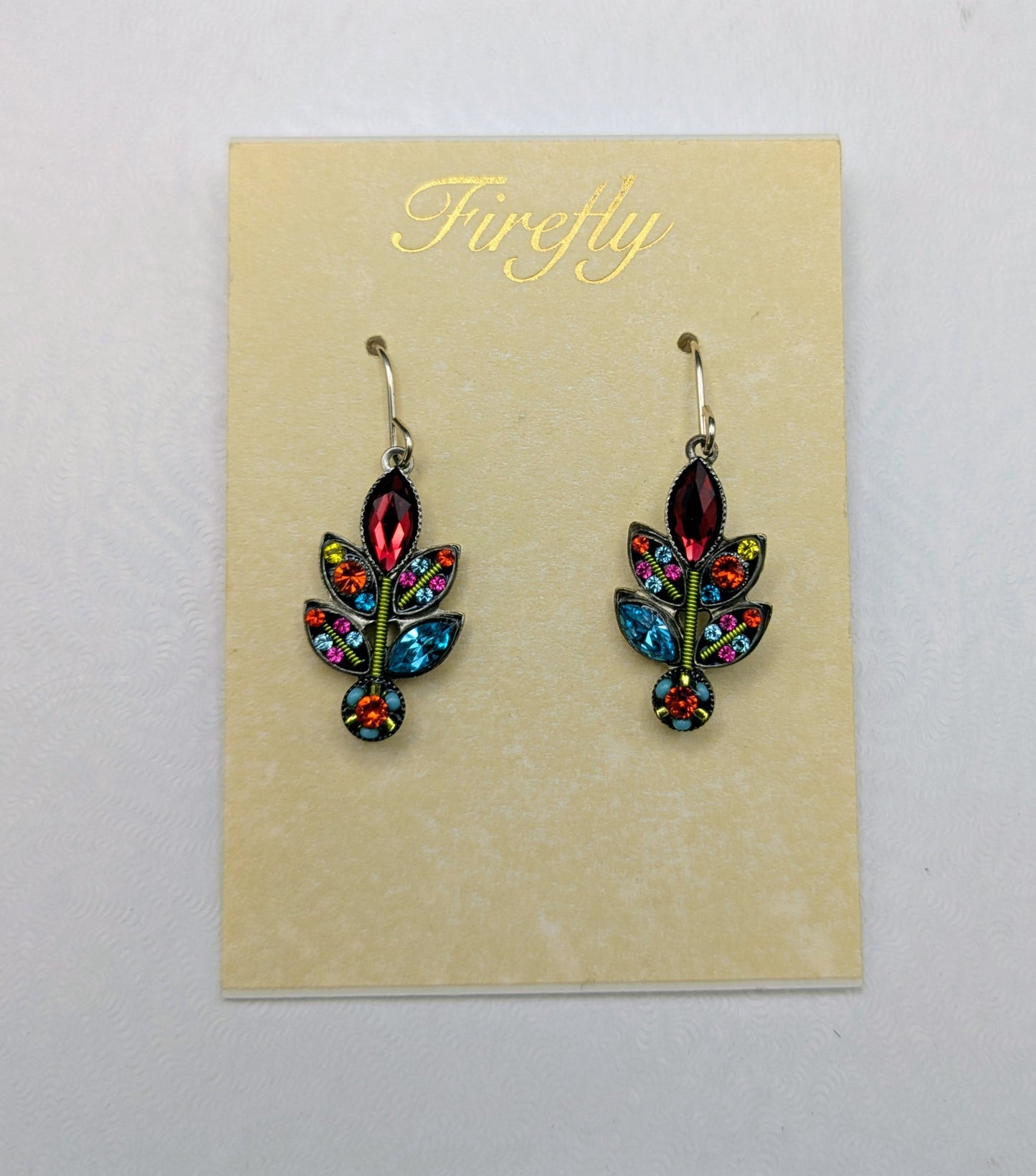 A pair of colorful dangling earrings displayed on a cream card labeled 'Firefly' inside a white box. The earrings feature a leaf-like design with red, blue, green, and orange enamel details and gold-tone hooks.