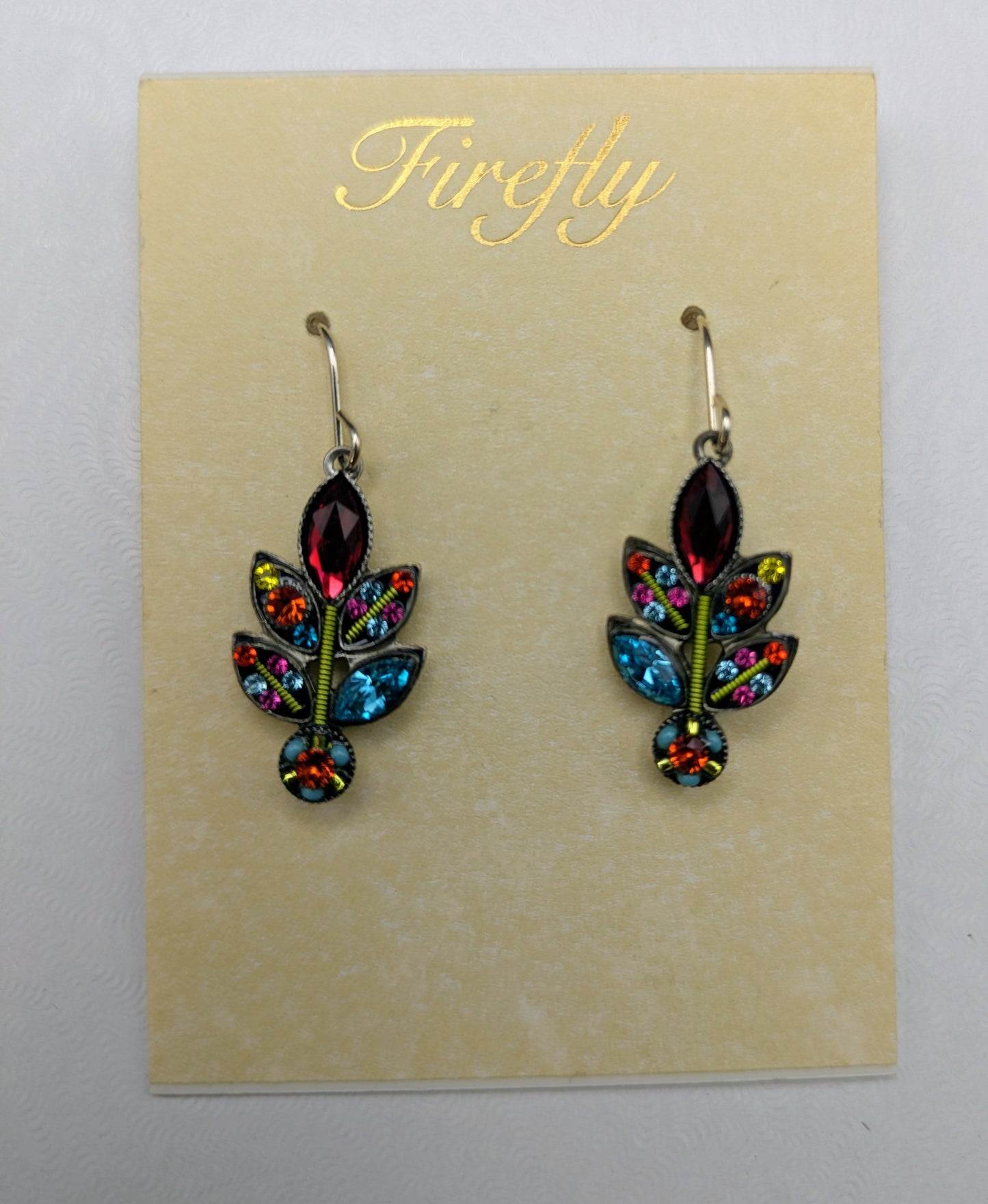 A pair of dangling leaf-shaped earrings displayed on a cream card printed with the brand name 'Firefly'. The earrings feature iridescent multicolor rhinestones in green, blue, and orange, with dark purple marquise-cut stones at the top and bottom, attached to silver hooks.