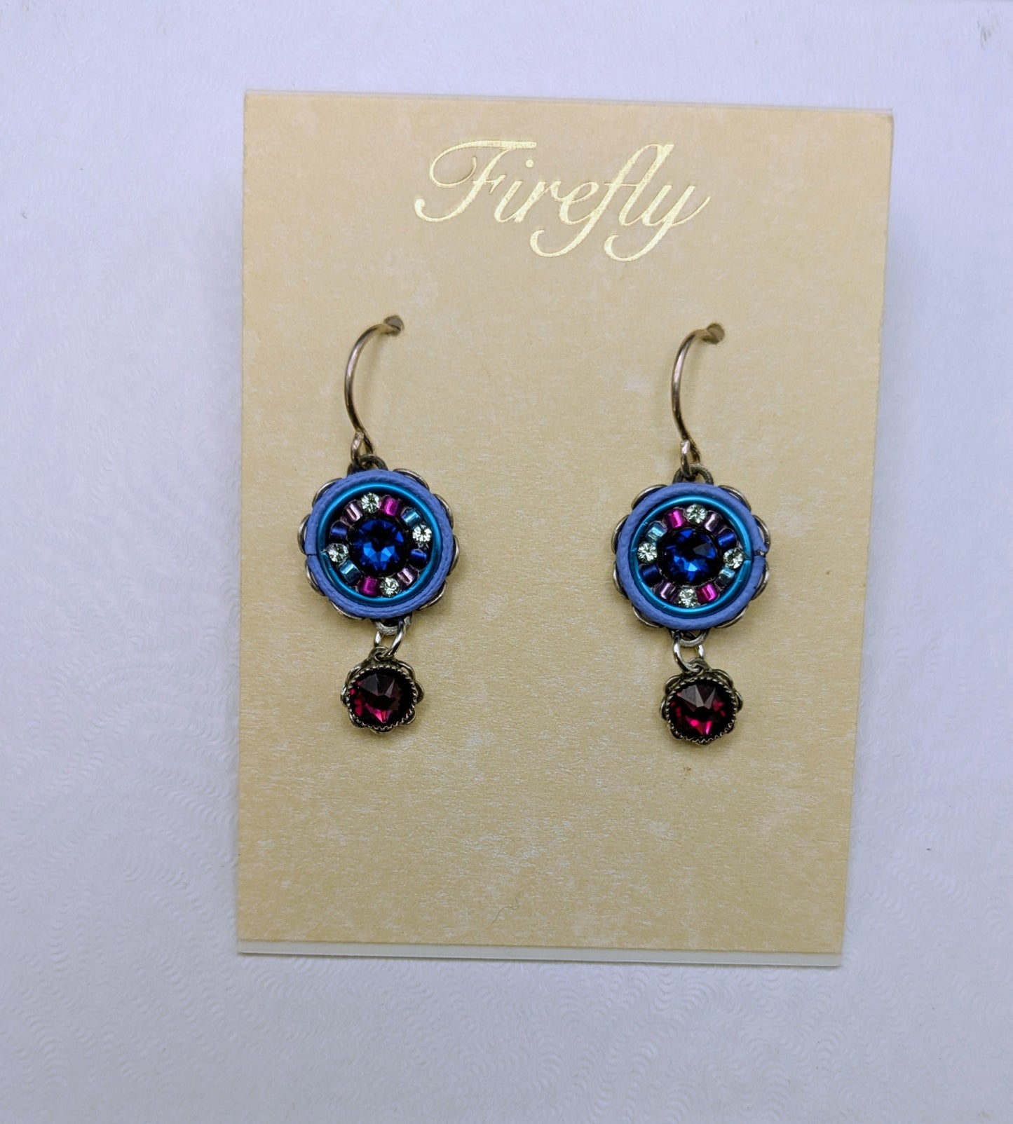 A pair of handmade dangle earrings displayed on a beige card labeled Firefly, resting on a larger white backing card against a neutral gray background.