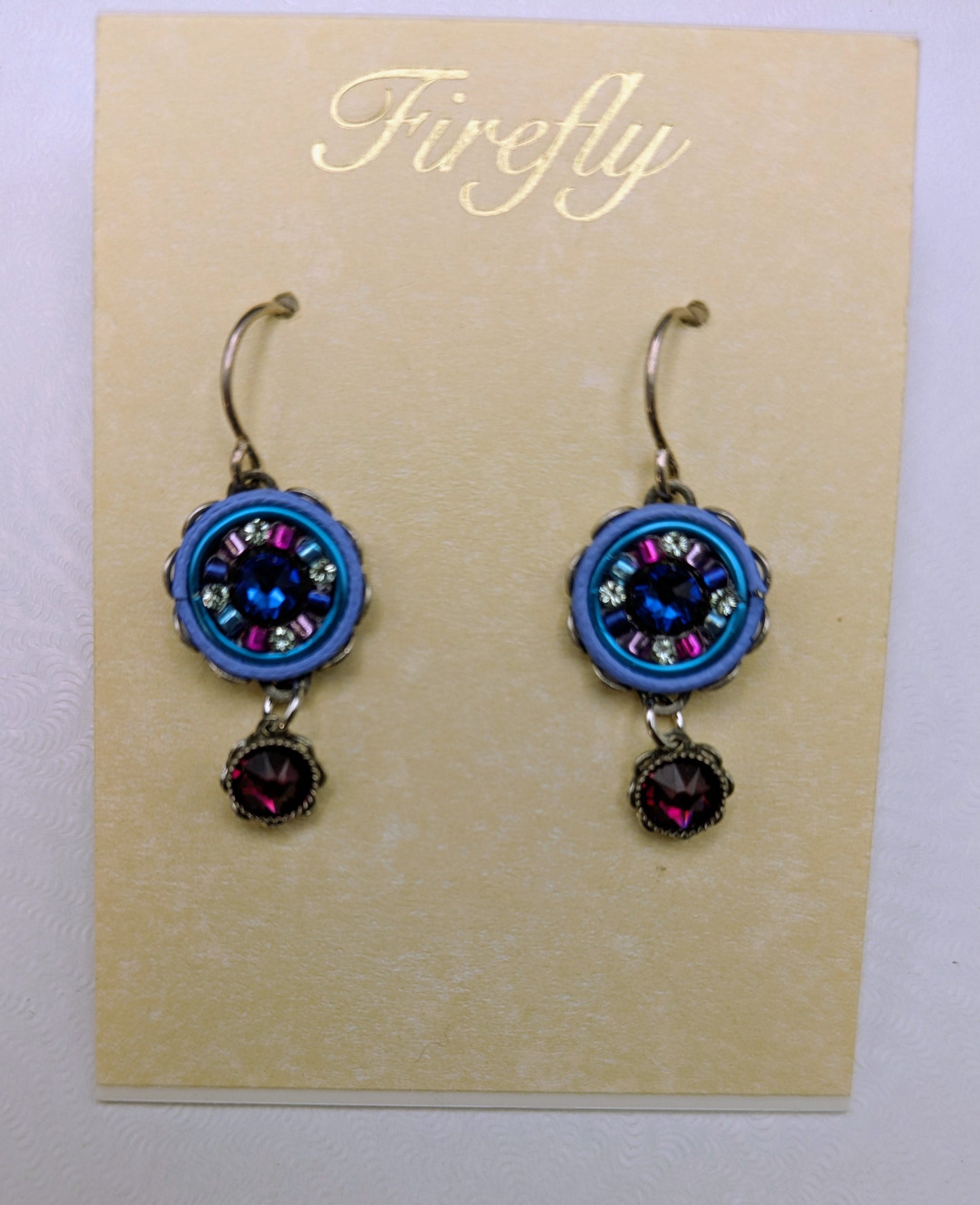 A pair of handmade dangle earrings displayed on a beige card. Each earring features a round blue and silver-toned beaded medallion with a dark blue center stone, a small dark pink gemstone drop, and a silver hook.