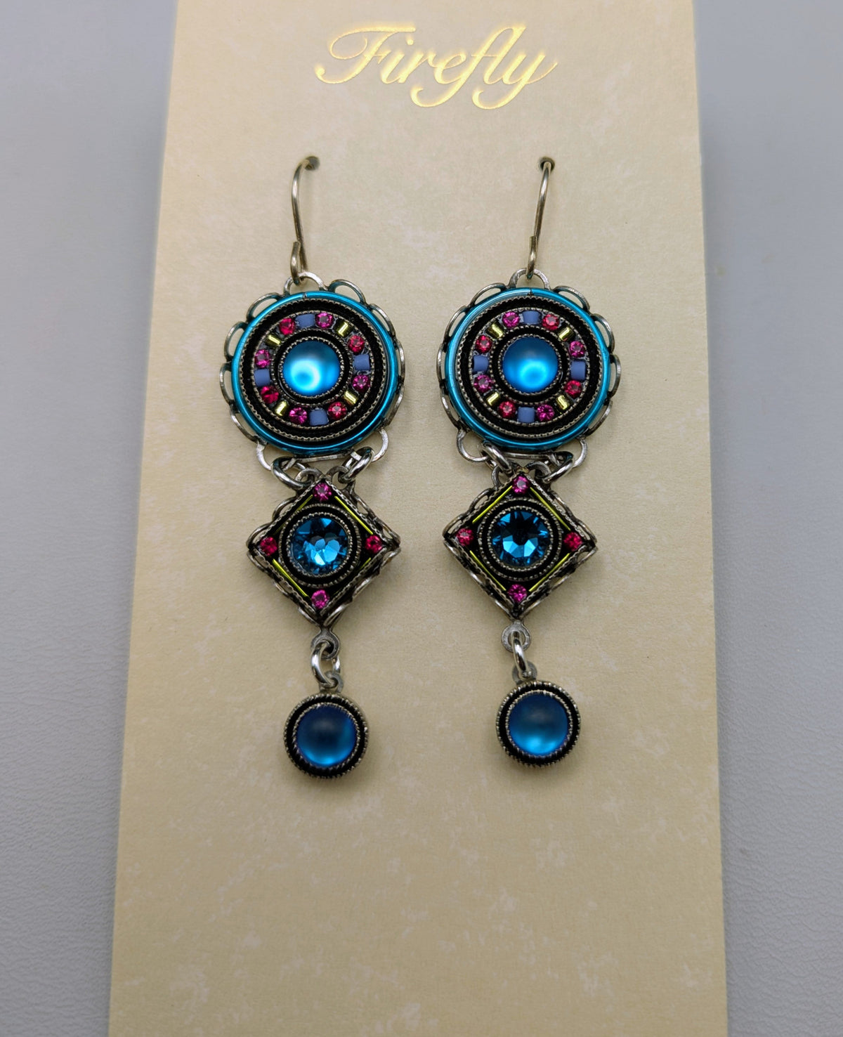 A pair of ornate drop earrings displayed on a cream-colored card. The earrings feature turquoise blue circular charms with pink and silver accents, connected by silver-toned chains to matching dangling charms with a blue teardrop bead at the bottom.