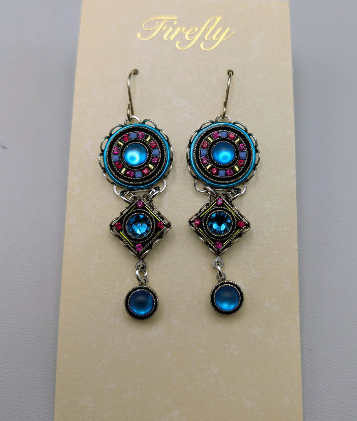A pair of ornate drop earrings displayed on a cream-colored card. The earrings feature turquoise blue circular charms with pink and silver accents, connected by silver-toned chains to matching dangling charms with a blue teardrop bead at the bottom.