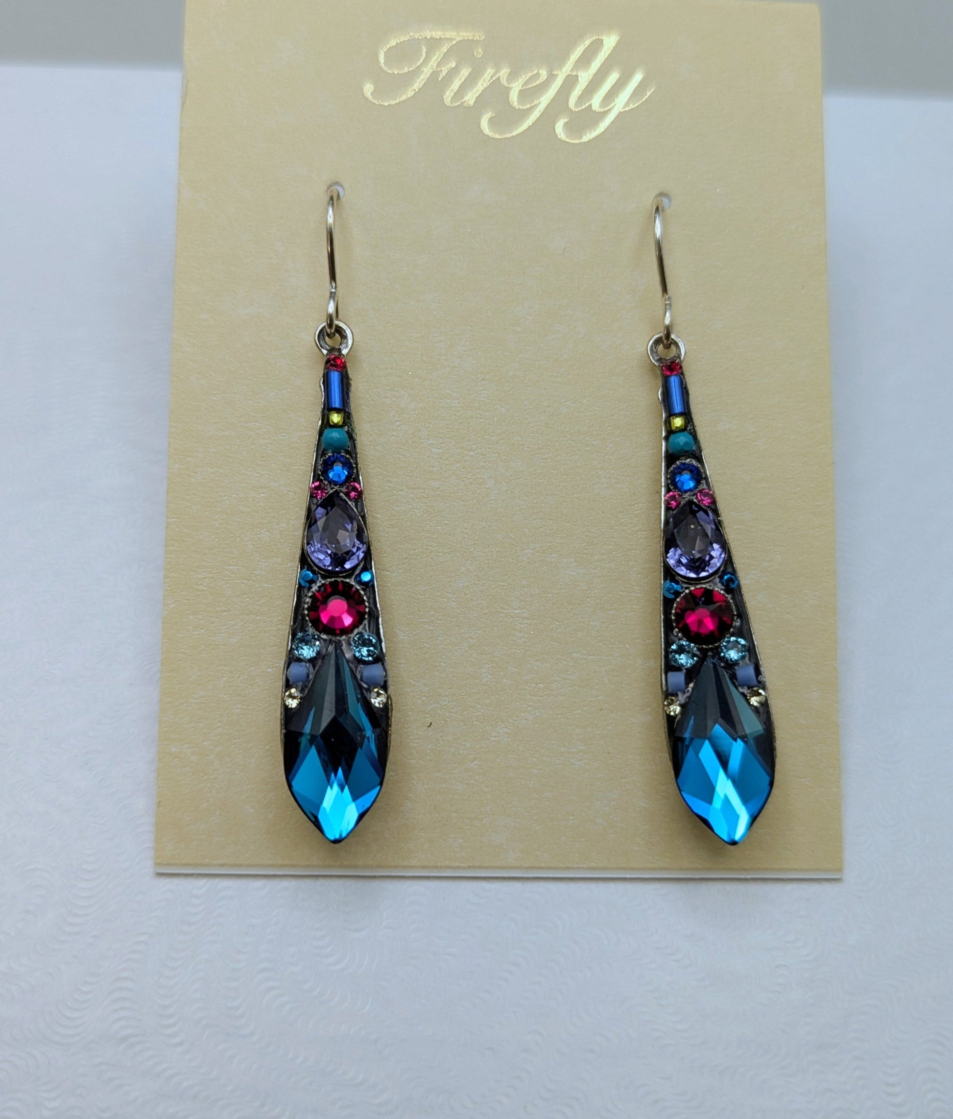 A pair of long teardrop-shaped dangling earrings displayed on a beige card inside a white jewelry box. The earrings have silver-toned metal settings with faceted blue, purple, and pink stones.