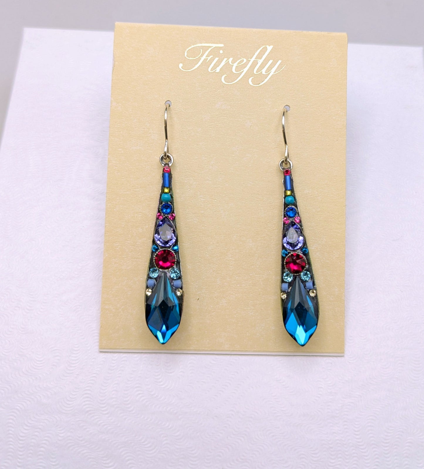 A pair of long teardrop-shaped dangling earrings displayed on a beige card labeled 'Firefly' inside a white presentation box. The earrings have a blue base with colorful floral details in red, pink, purple, and green, and are suspended from silver hooks.