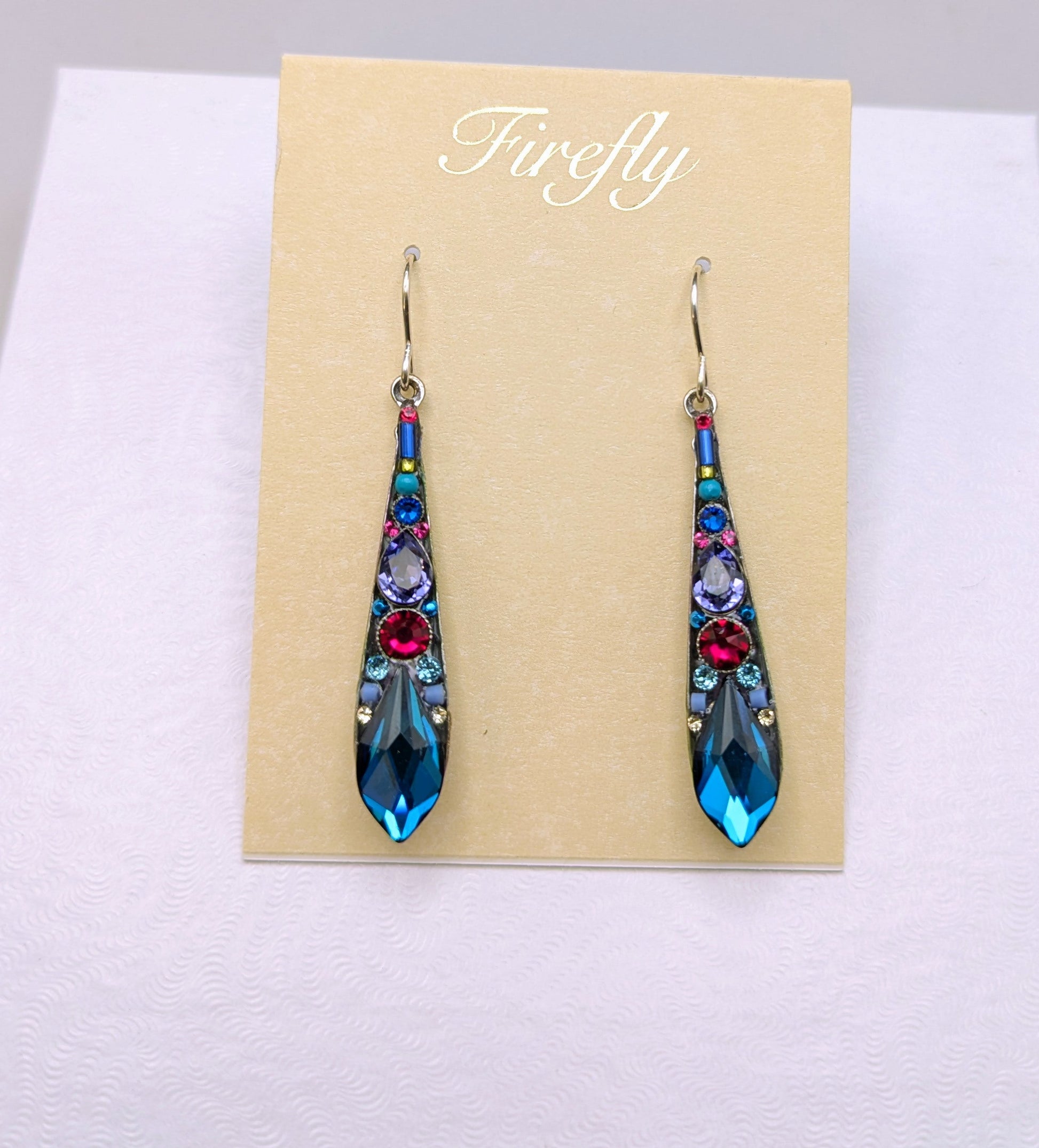 A pair of long teardrop-shaped dangling earrings displayed on a beige card labeled 'Firefly' inside a white presentation box. The earrings have a blue base with colorful floral details in red, pink, purple, and green, and are suspended from silver hooks.