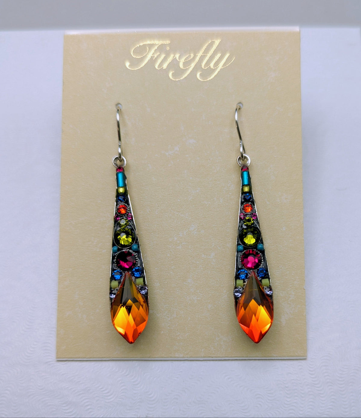 A pair of Firefly brand teardrop-shaped dangling earrings with colorful rhinestones and a bright orange flame-shaped crystal at the bottom, displayed on a beige card inside a white box.
