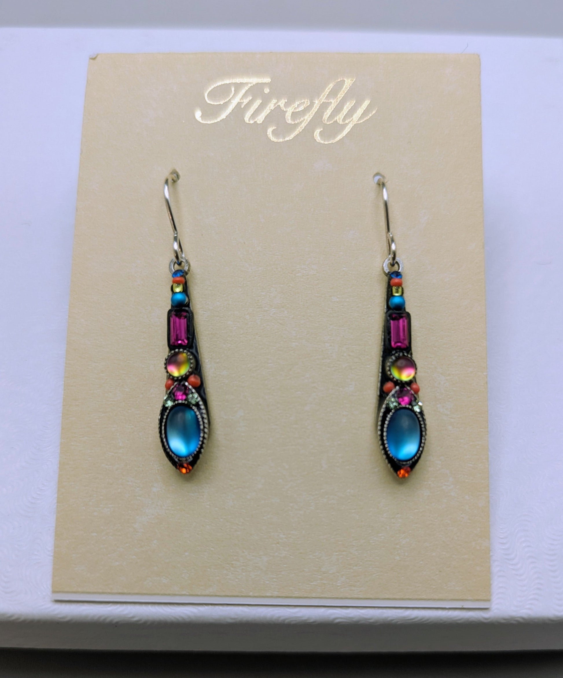 A pair of dangling earrings displayed on a beige card labeled 'Firefly' inside a white box. The earrings have silver-toned hooks and settings with small iridescent yellow and pink beads, a magenta rectangular stone, and a larger blue oval stone at the bottom.